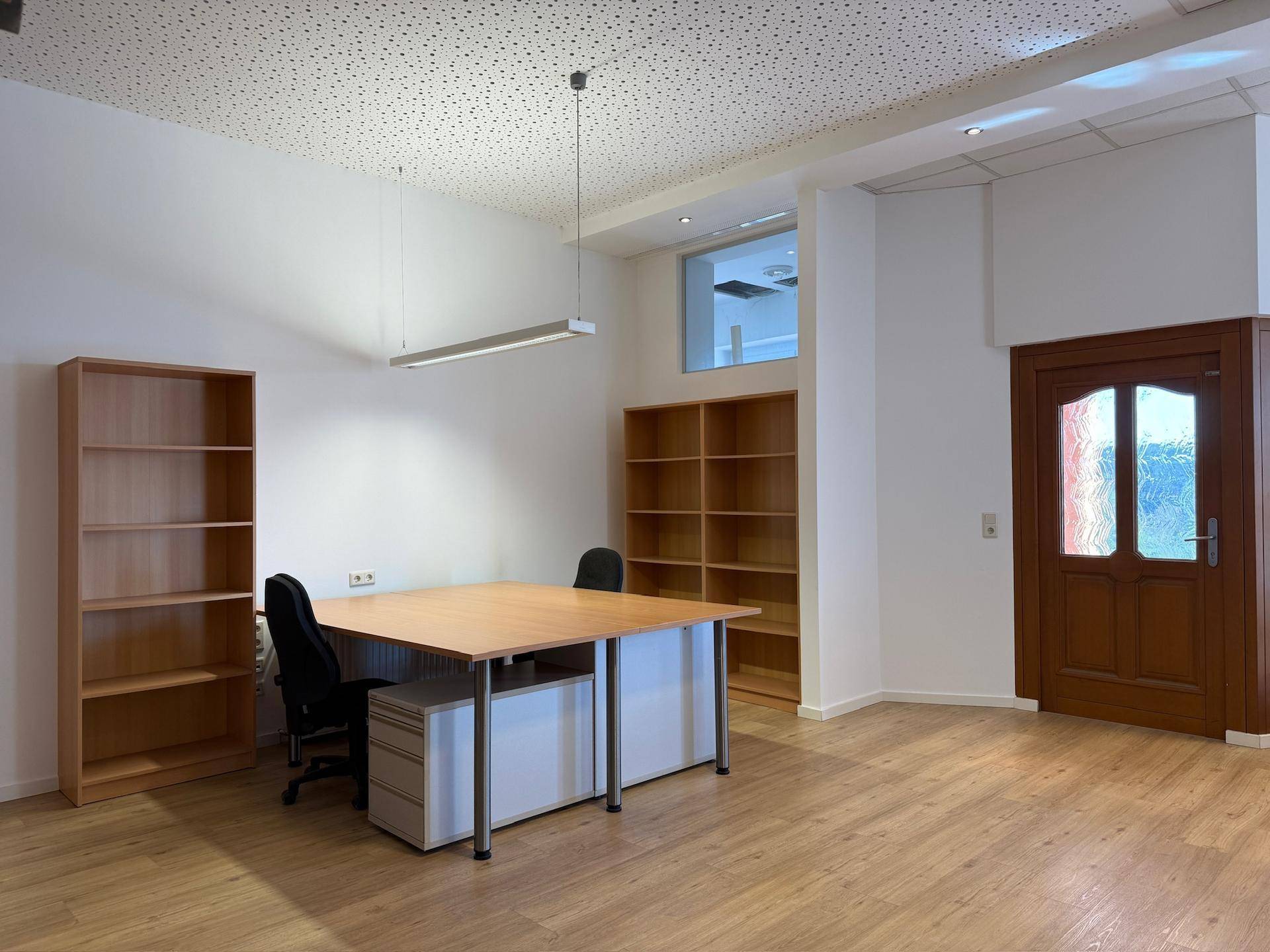 Bürofläche mit hohen Decken, Laminatboden und Holzregalen, ausgestattet mit einem Arbeitstisch.