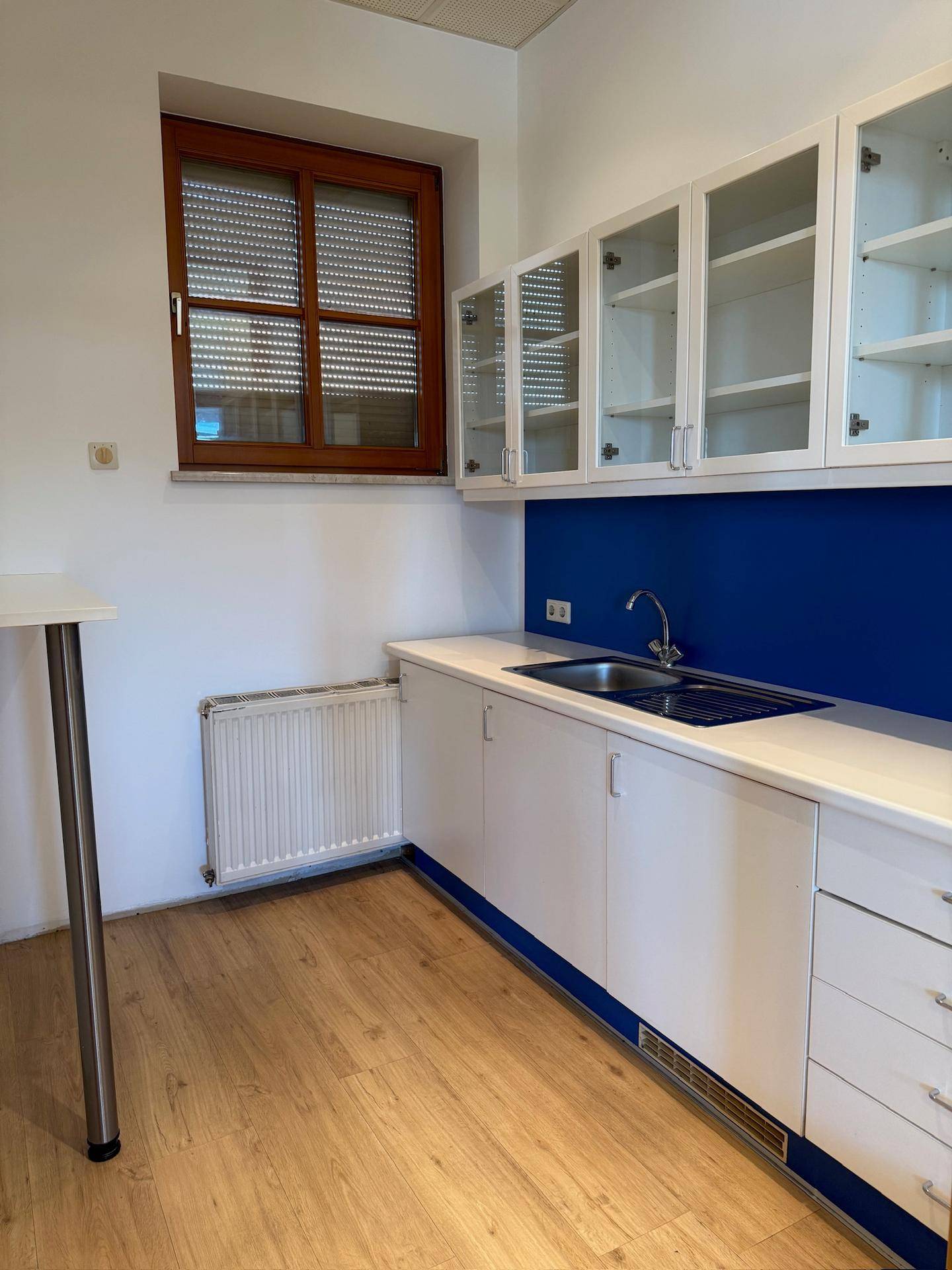Kleine Küchenzeile mit weißen Schränken, blauer Rückwand und einem Fenster mit Jalousien.