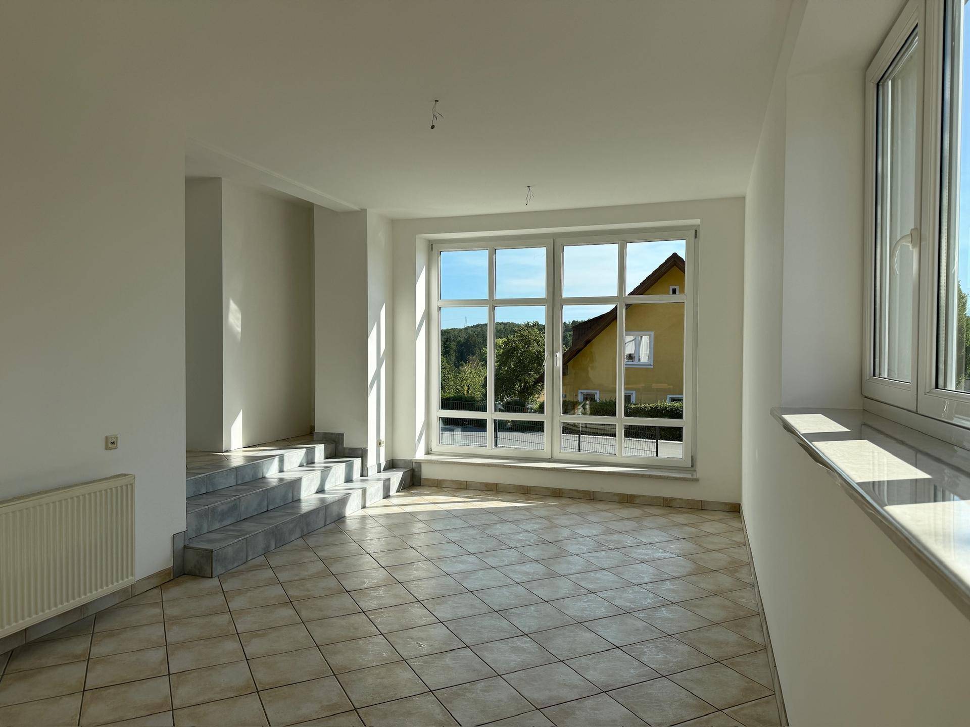 Heller Raum mit Fliesenboden, großen Fenstern und einer Treppe, die zu einem anderen Bereich führt.