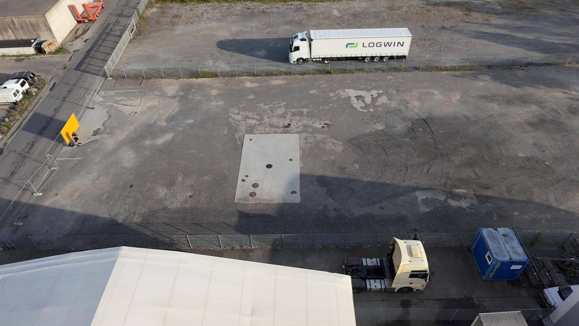 Detaillierte Luftansicht eines Industrieareals mit Lastwagen, umzäuntem Gelände und Betonplatten auf dem Boden.