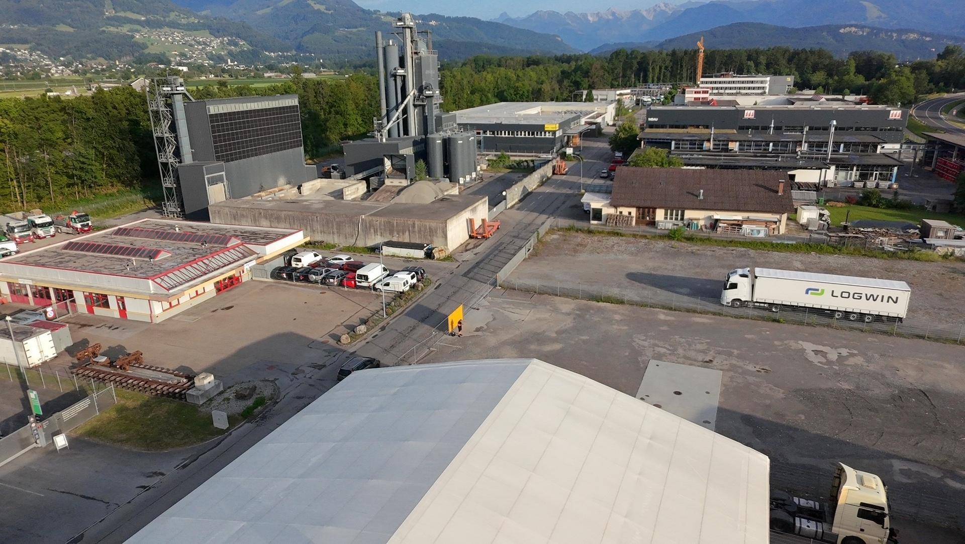 Weitläufige Luftaufnahme eines Industriegebiets mit verschiedenen Gebäuden, Lastwagen und Bergen im Hintergrund.