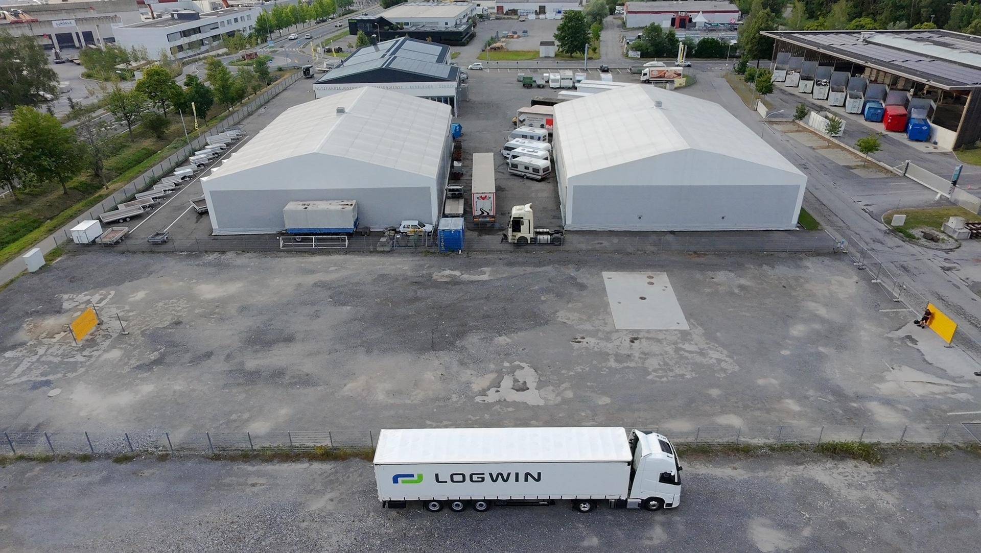 Luftbild eines Industrieareals mit zwei großen, hellen Lagerhallen, Lastwagen und einer weitläufigen Freifläche.