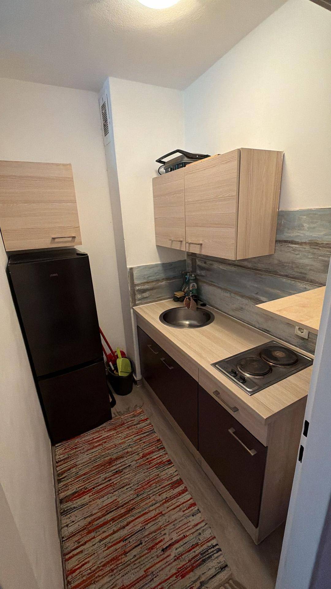 Kleine Küchenzeile mit hellen Holzfronten, zwei Kochplatten und einem Kühlschrank.