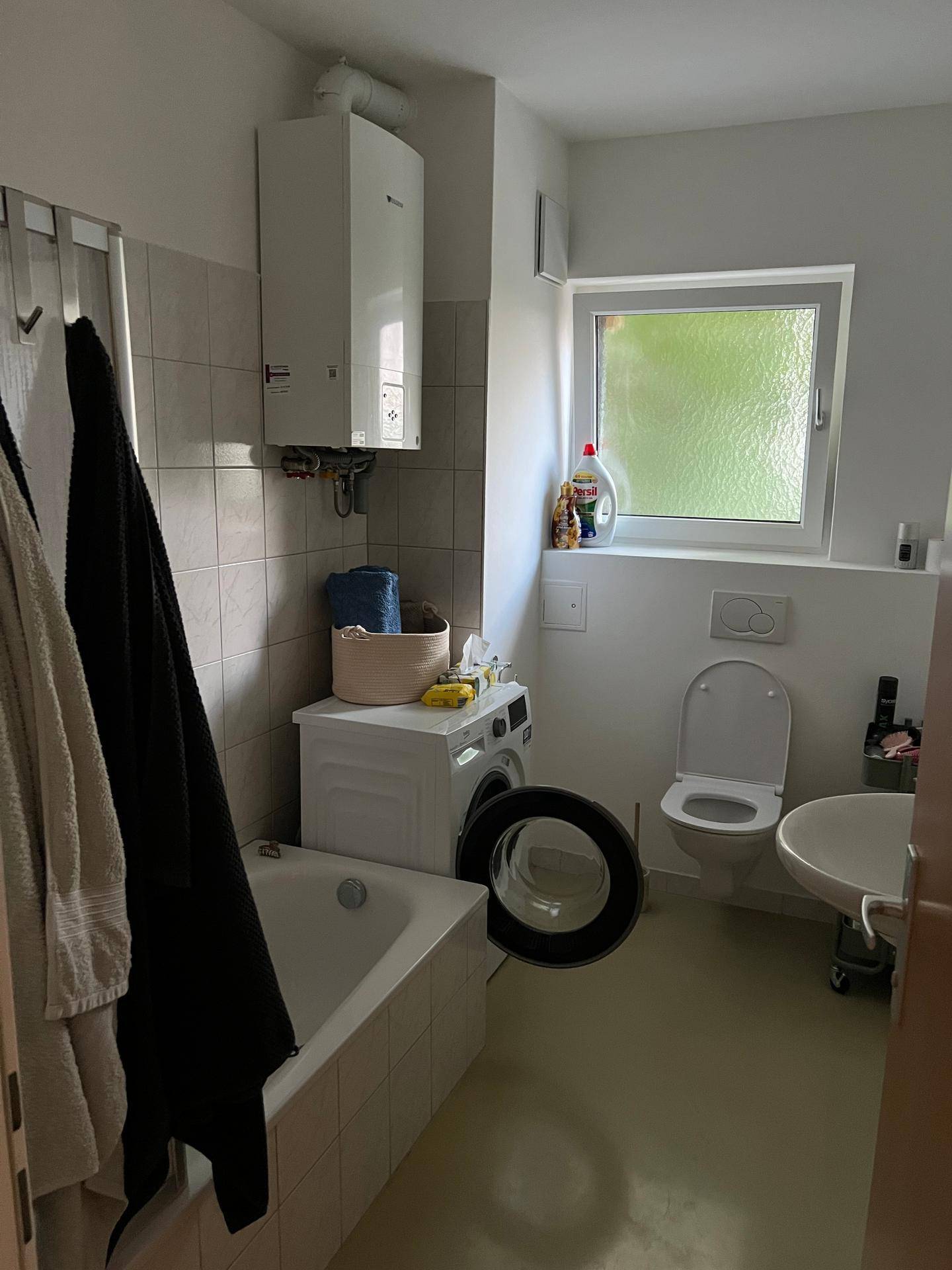 Badezimmer mit Badewanne, Toilette, Waschbecken und Waschmaschine, hell gefliest und mit Fenster.