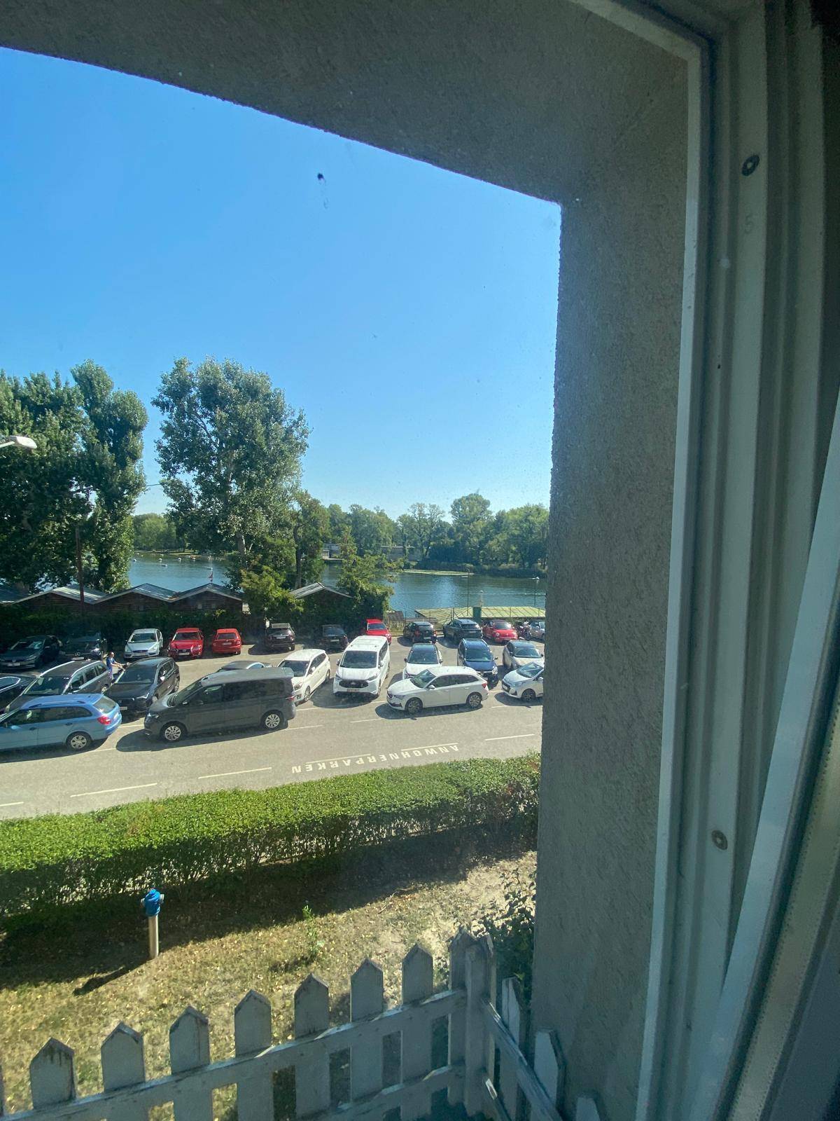 Blick aus dem Fenster auf einen Parkplatz, eine Straße und einen See mit Bäumen im Hintergrund.
