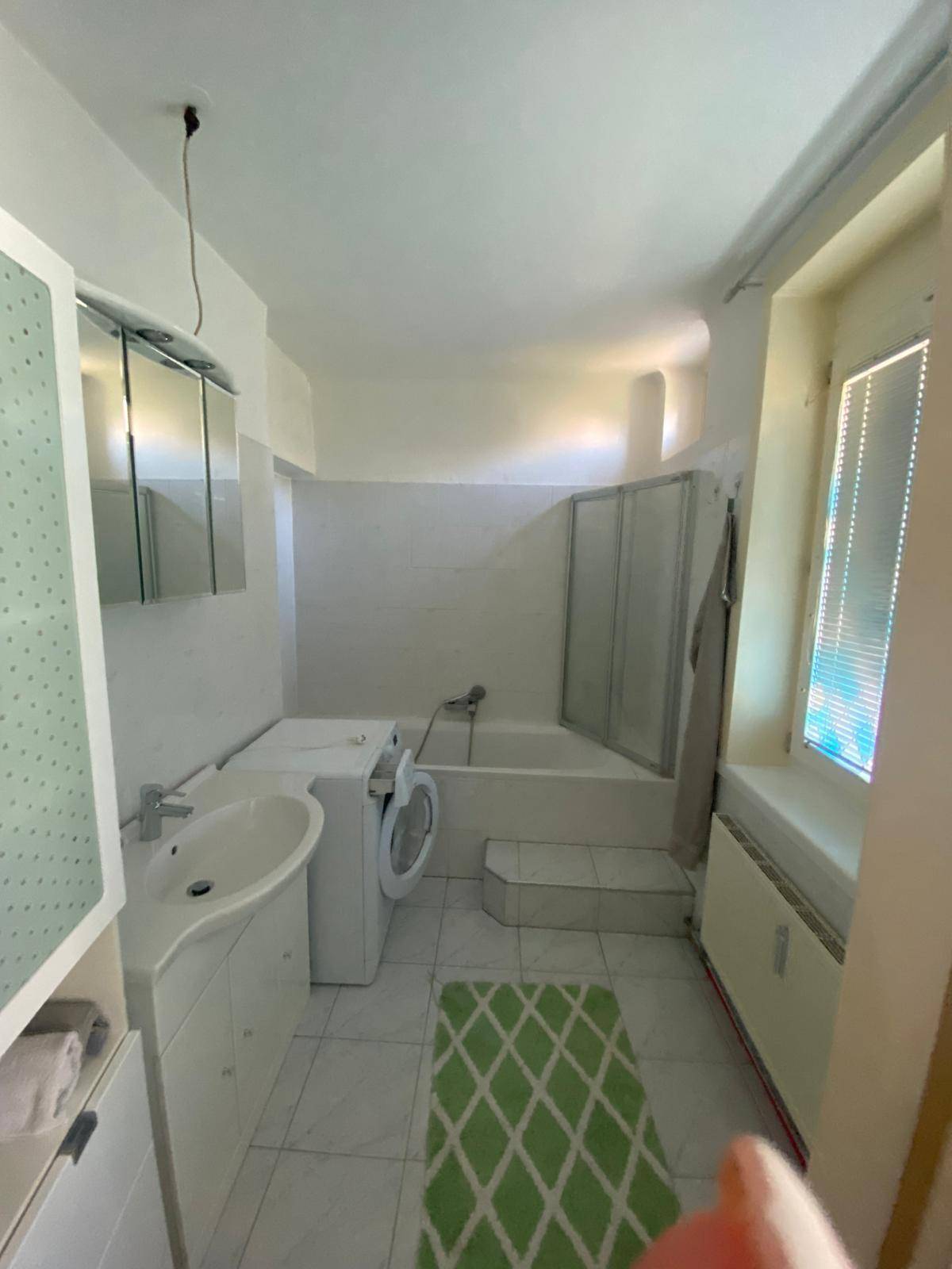 Helles Badezimmer mit Badewanne, Duschabtrennung, Waschbecken und Waschmaschine sowie einem Fenster.