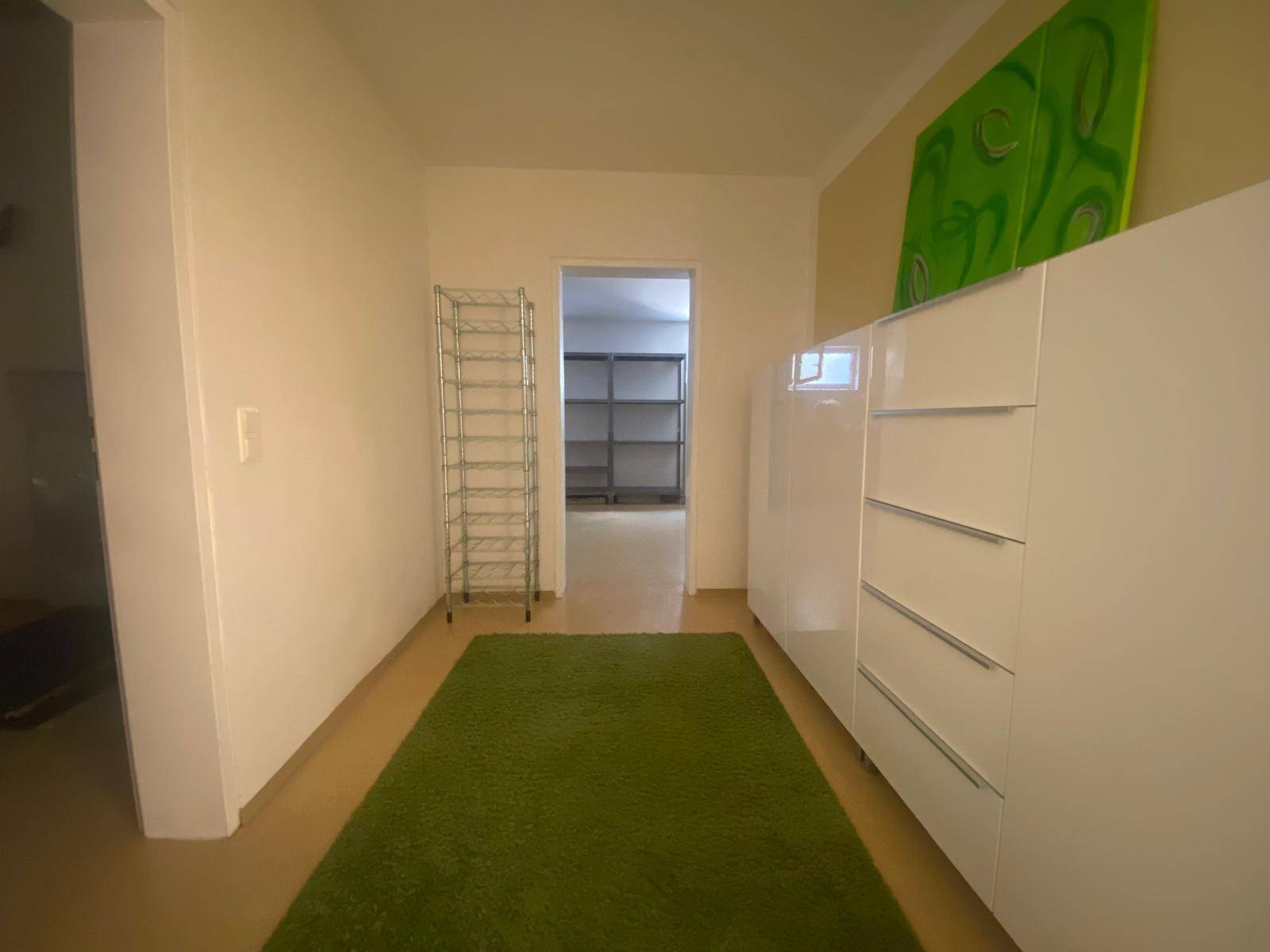 Heller Kellerflur mit weißer Kommode, grünem Teppich und Zugang zu weiteren Räumen.