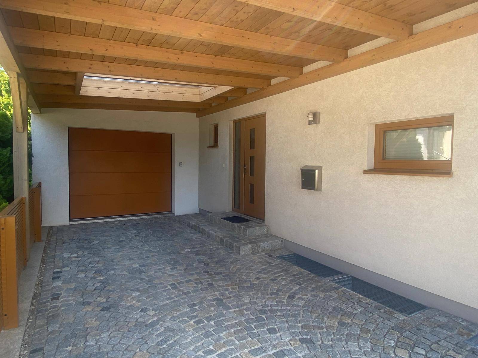 Überdachter Carport mit Holzbalken, gepflastertem Boden und Garagentor.