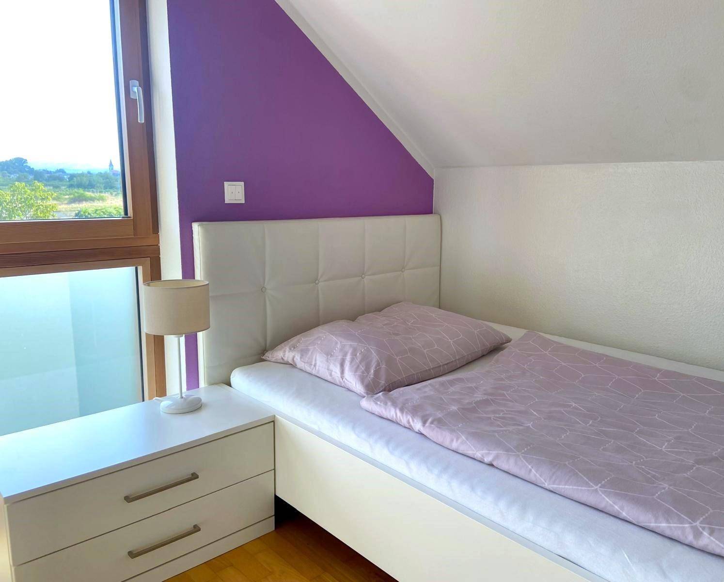 Helles Kinderzimmer mit Einzelbett, lila Wandakzent und Blick ins Grüne.