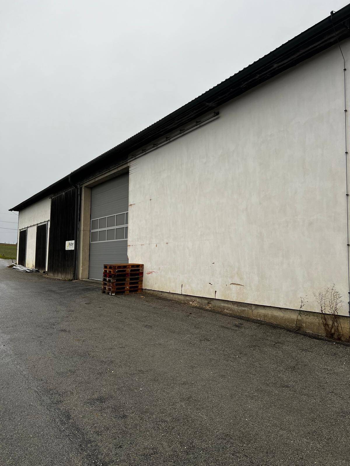 Seitenansicht des Gebäudes mit großem grauen Garagentor und weißer Fassade, davor gestapelte Paletten.