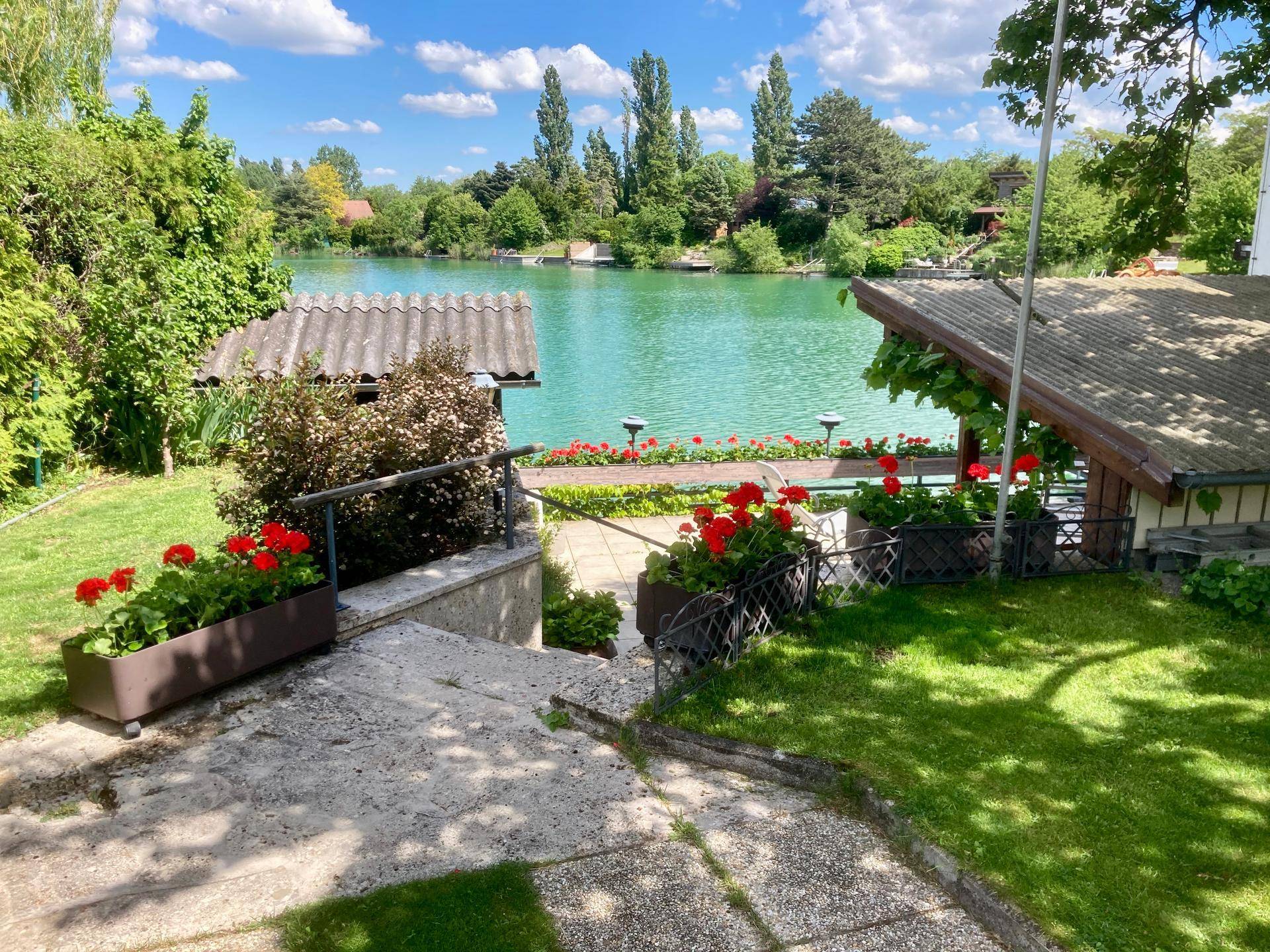 Grüner Gartenbereich mit Treppe zum See und blühenden Geranien unter blauem Himmel.