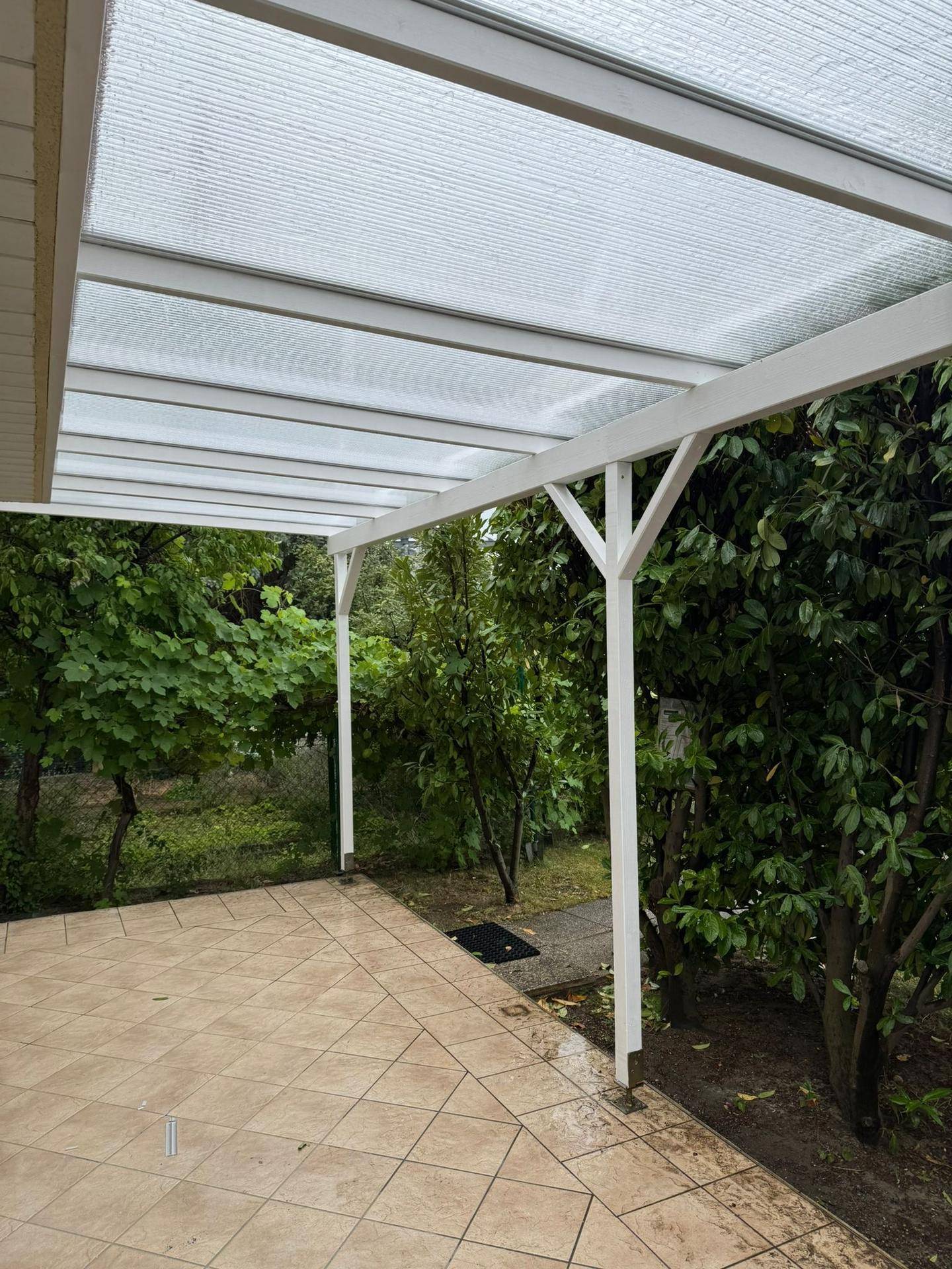 Überdachte Terrasse mit Fliesenboden und transparentem Dach, umgeben von Bäumen und Grün.