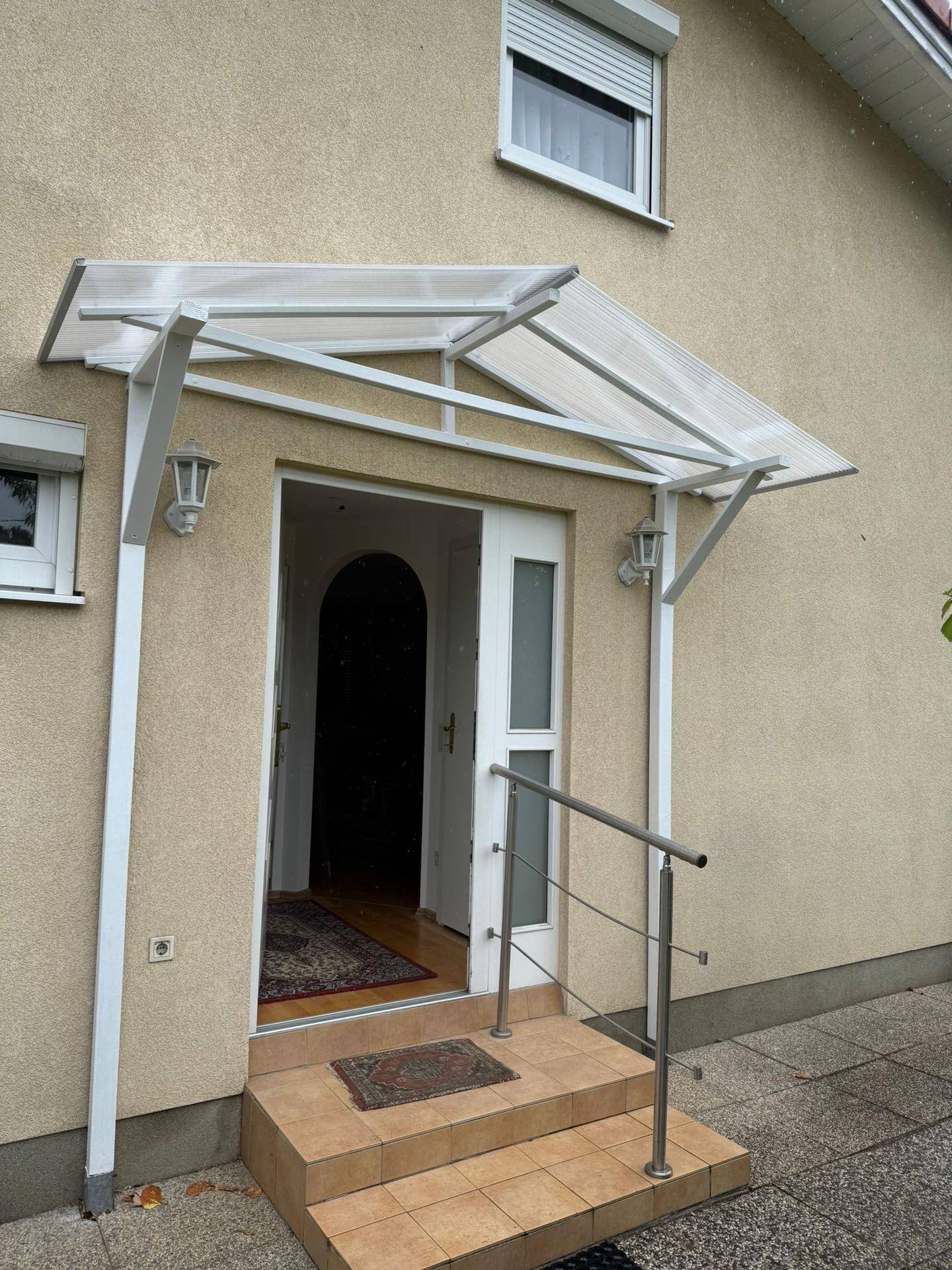 Hauseingang mit Treppe, Geländer und einem transparenten Vordach, beleuchtet von Wandlampen.