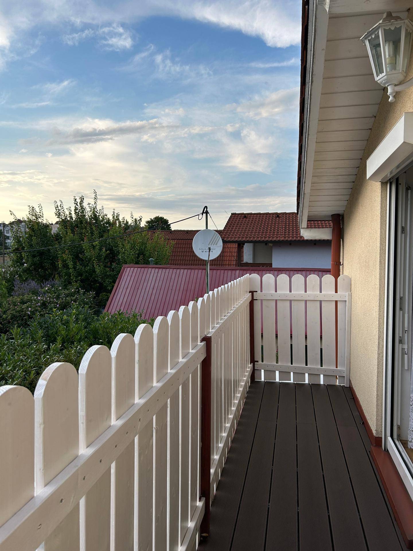 Kleiner Balkon mit dunklem Boden und weißem Lattenzaun, bietet Blick über Dächer und Bäume.