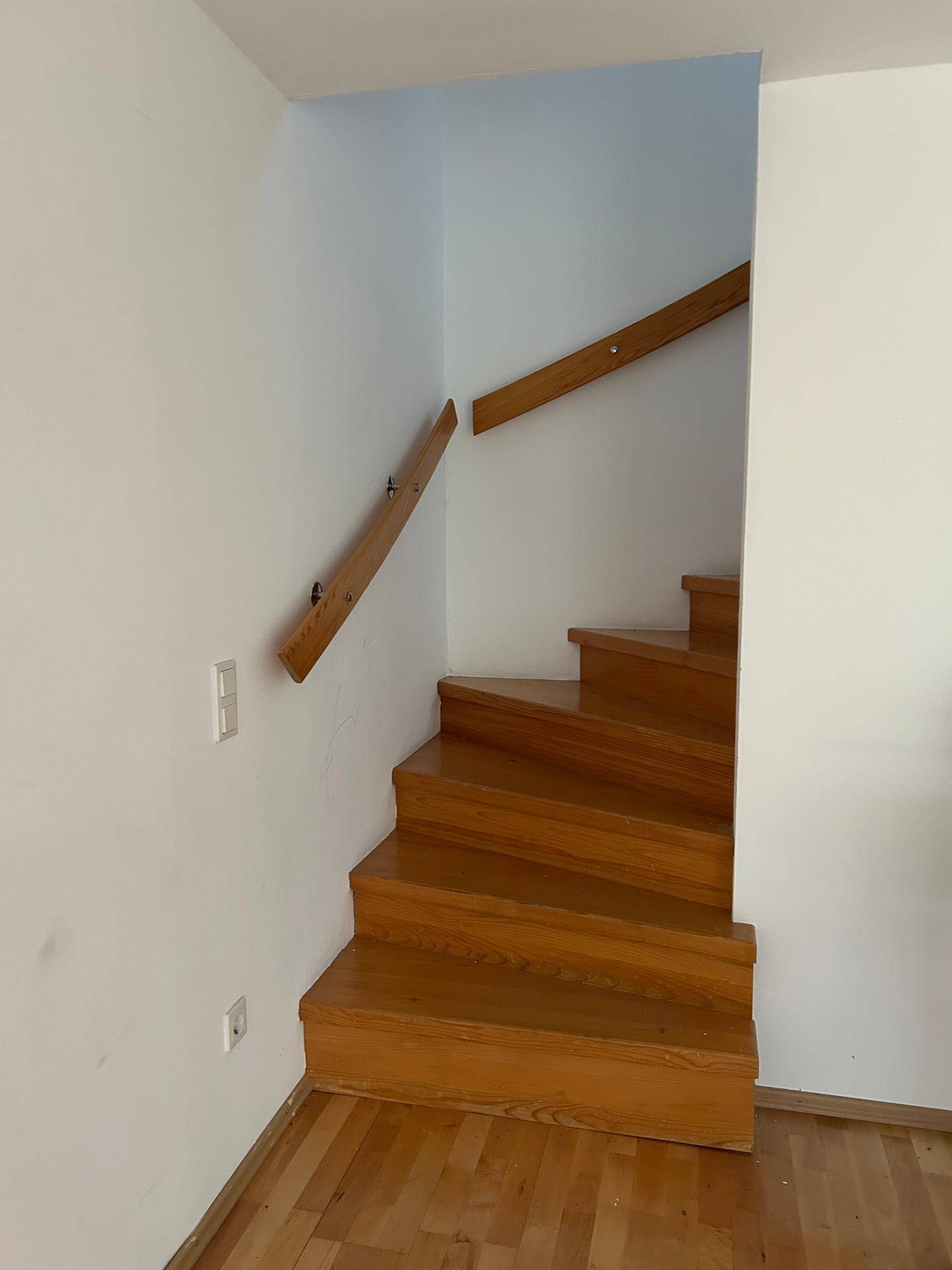 Holztreppe mit schlichtem Holzgeländer führt in die obere Etage, umgeben von weißen Wänden.