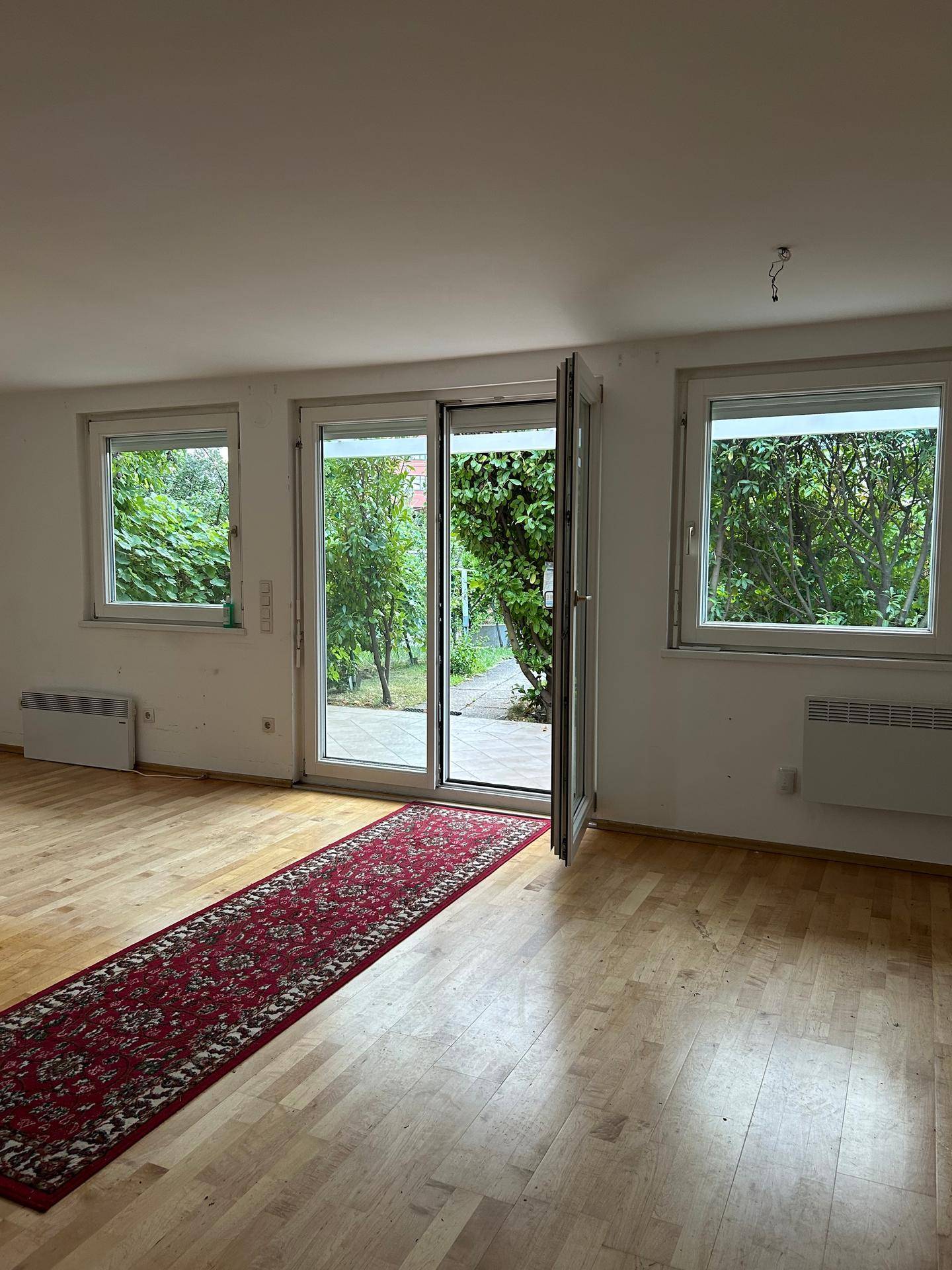 Leeres Wohnzimmer mit Holzboden, Heizkörpern und einer großen Glastür zum Garten.