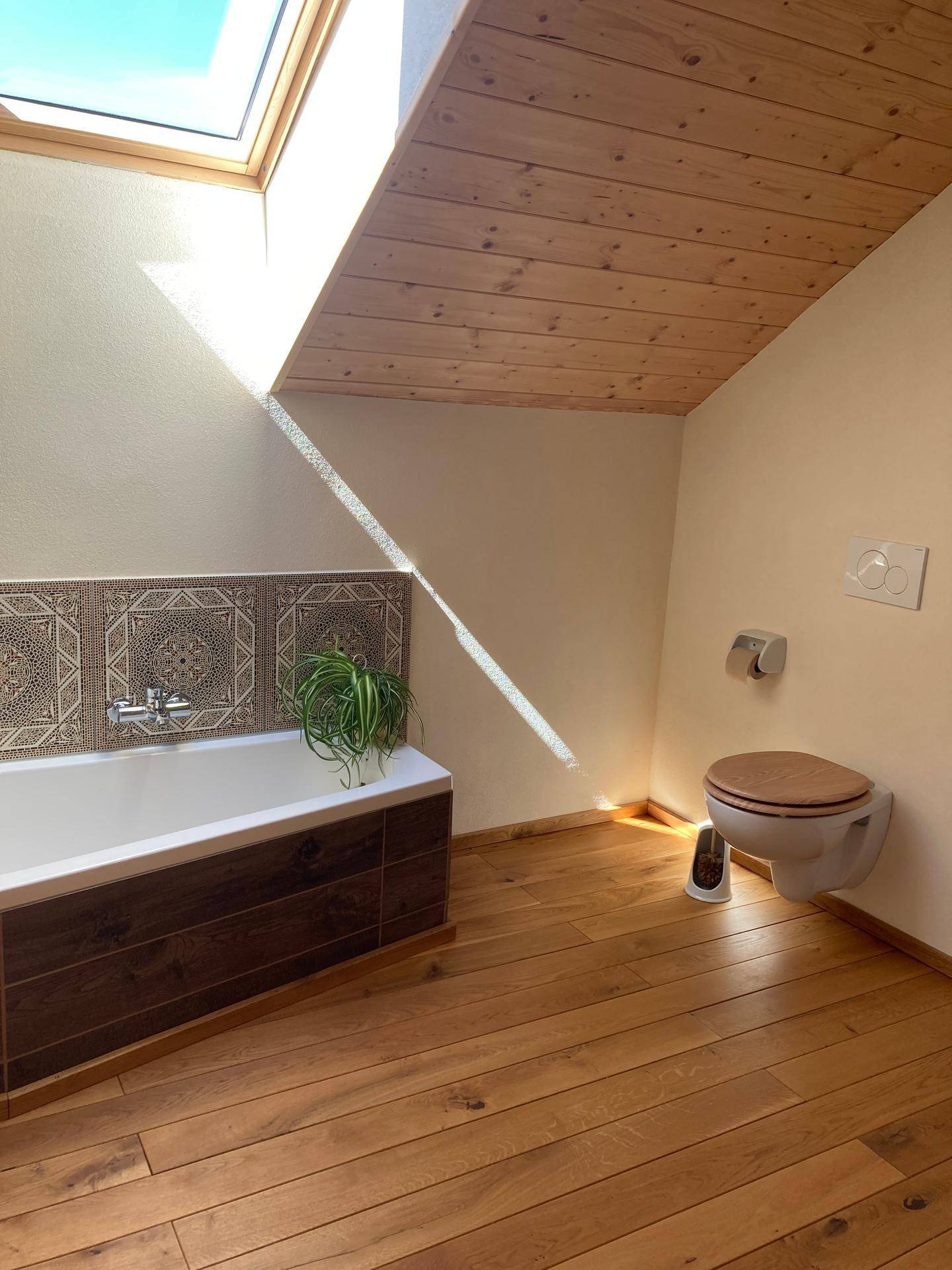Lichtdurchflutetes Badezimmer mit Badewanne, Toilette und Holzboden unter einem Dachfenster.