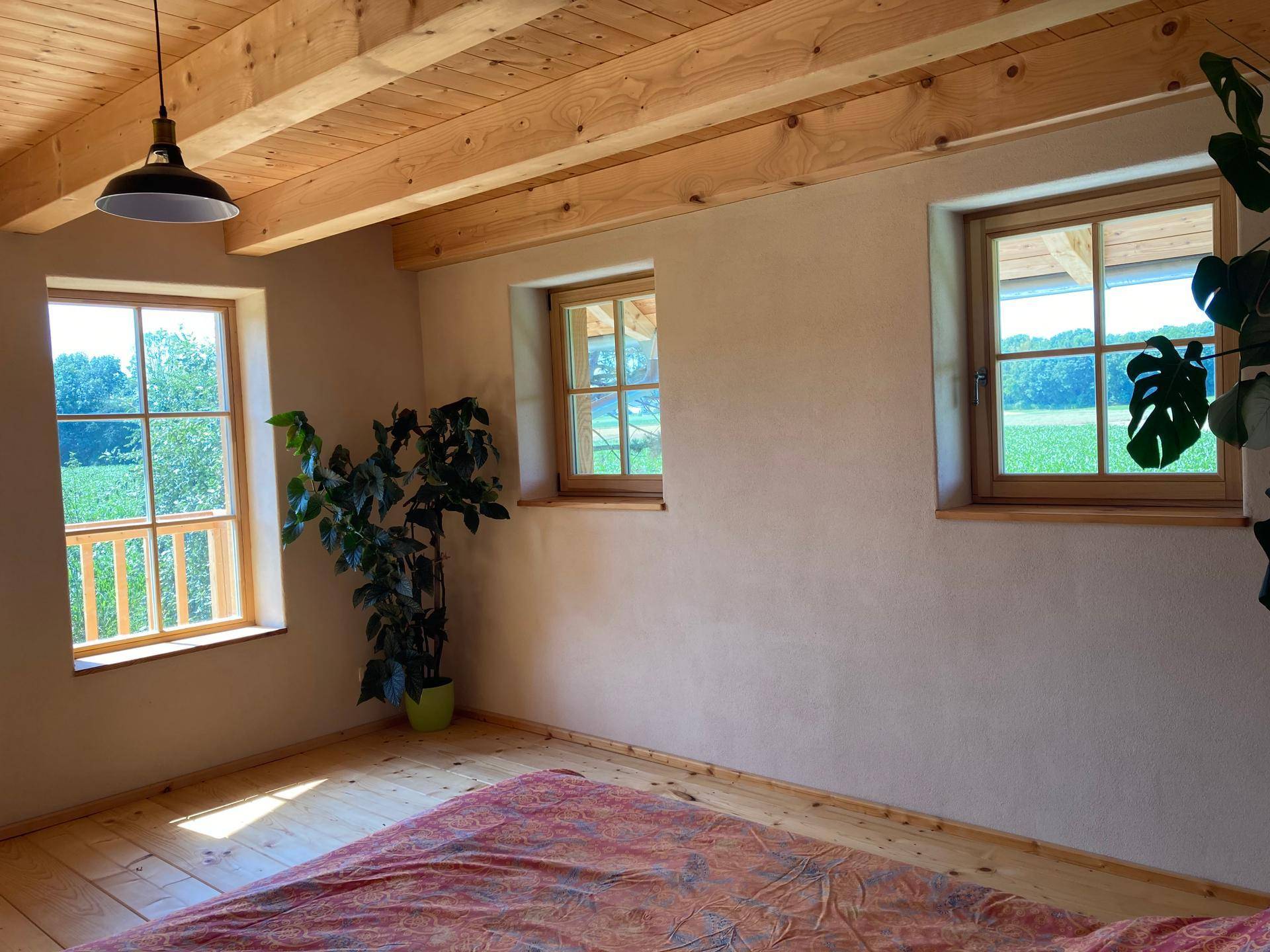 Helles Schlafzimmer mit Holzdielenboden, Deckenbalken und Fenstern mit Blick ins Grüne.