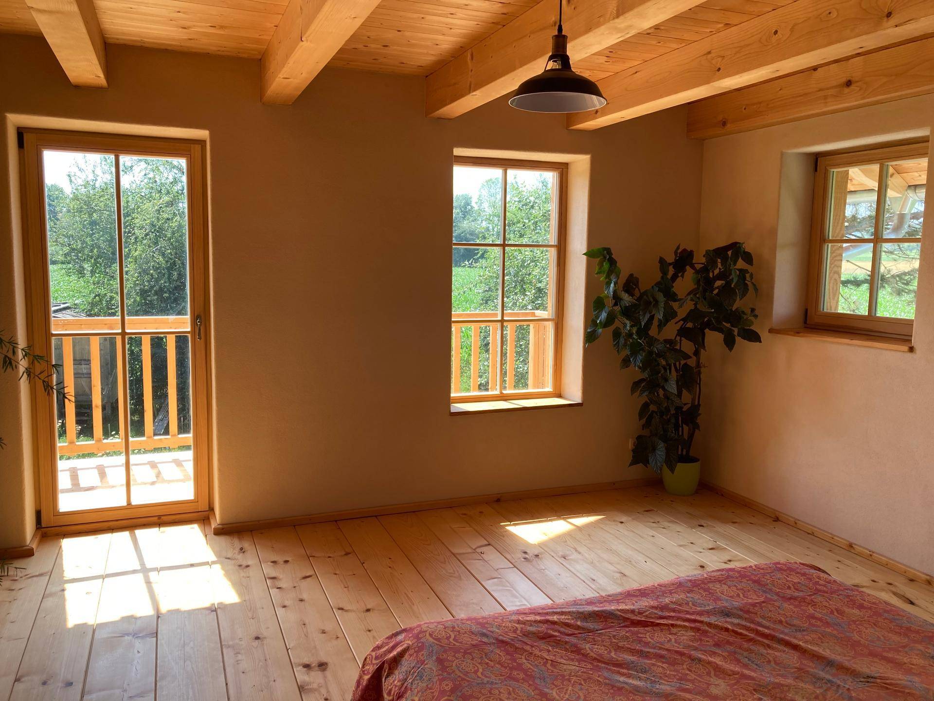 Schlafzimmer mit Holzdielenboden, Deckenbalken und Fenstern mit Blick ins Grüne.