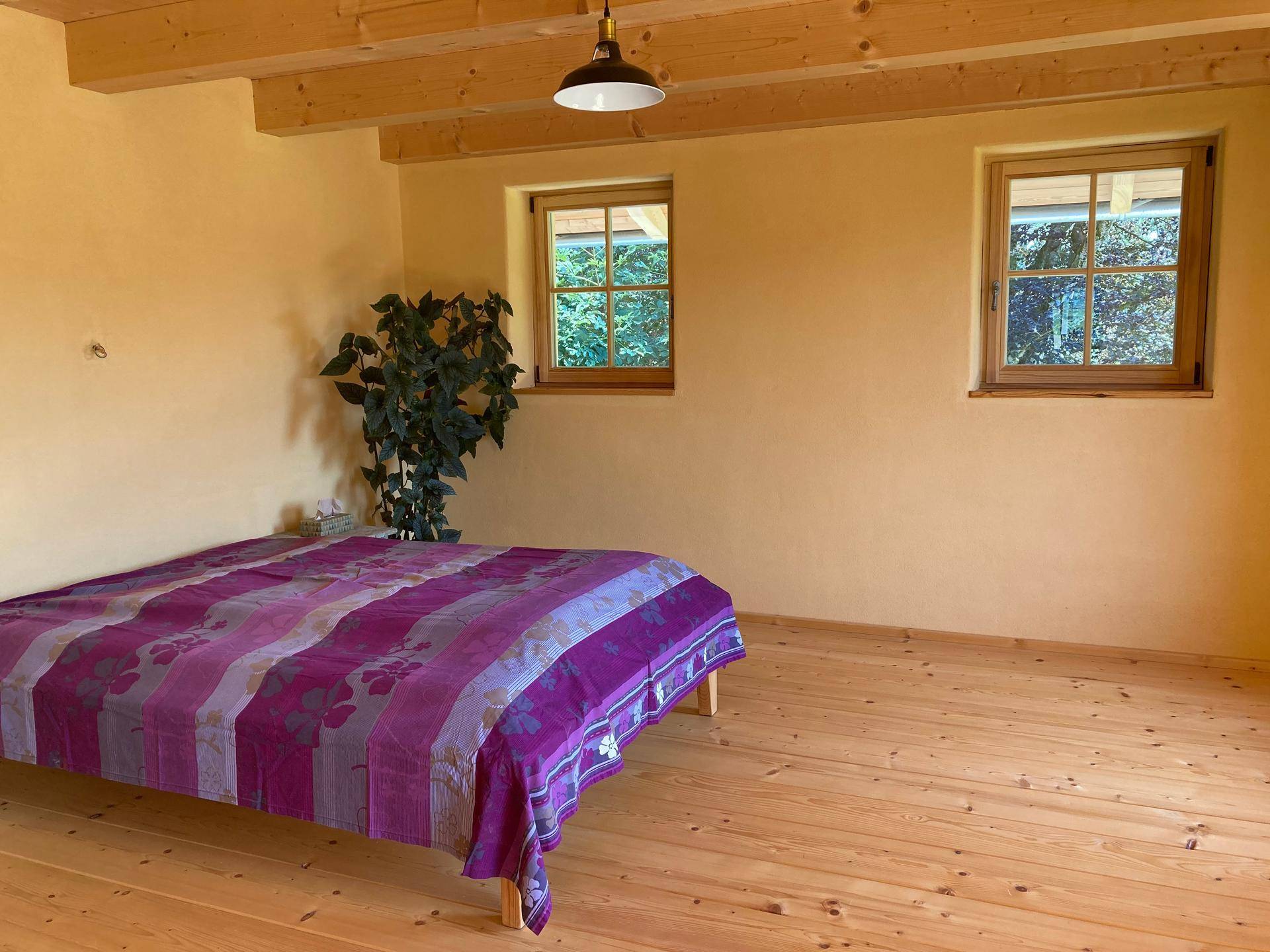 Schlafzimmer mit Holzdielenboden, gelben Wänden und Fenstern mit Blick ins Grüne.