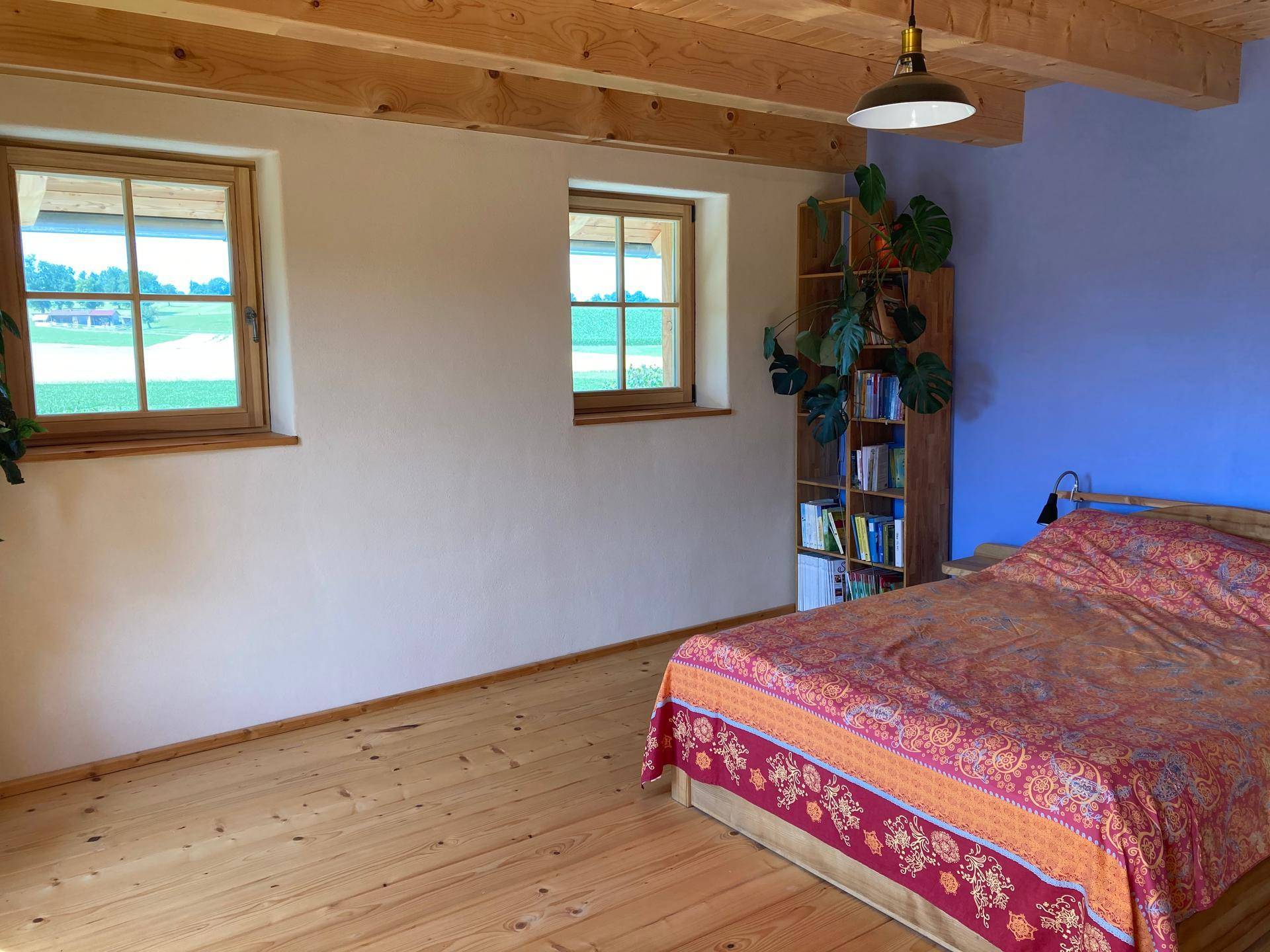 Schlafzimmer mit Holzdielenboden, Bett und Fenstern mit Blick auf die grüne Landschaft.
