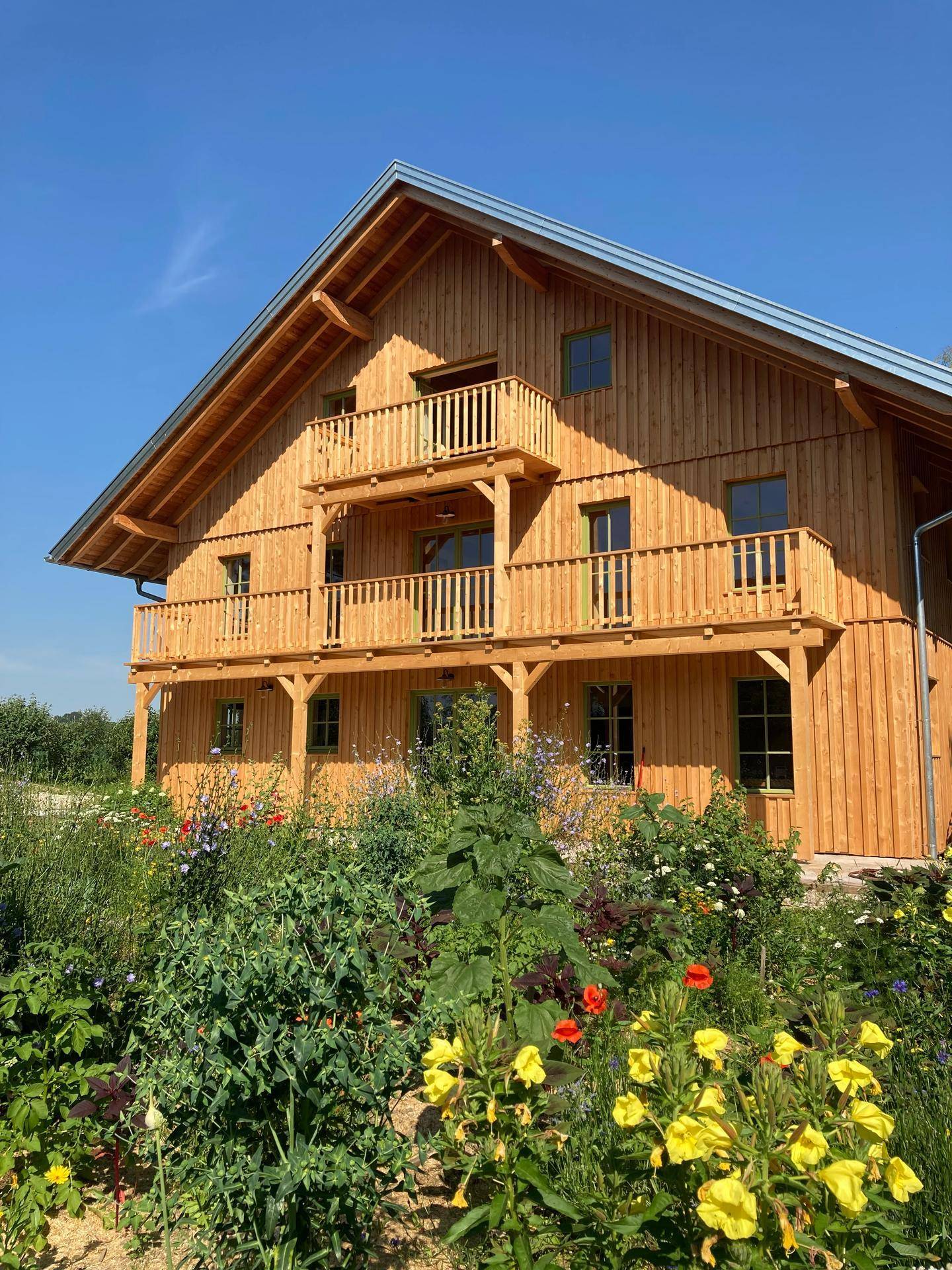 Holzhaus mit Balkonen und einem farbenfrohen Blumenbeet im Vorgarten unter blauem Himmel.