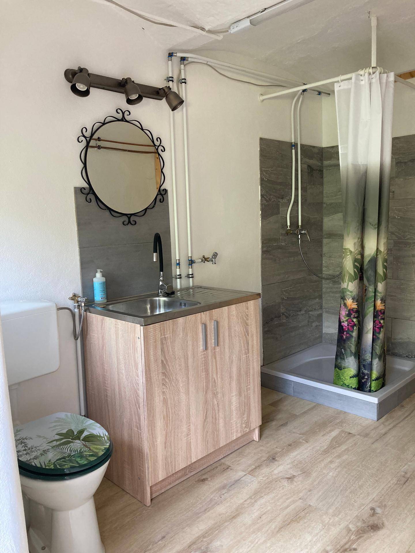 Einfaches Badezimmer mit Toilette, Waschbeckenunterschrank und einer Dusche mit floralem Vorhang.
