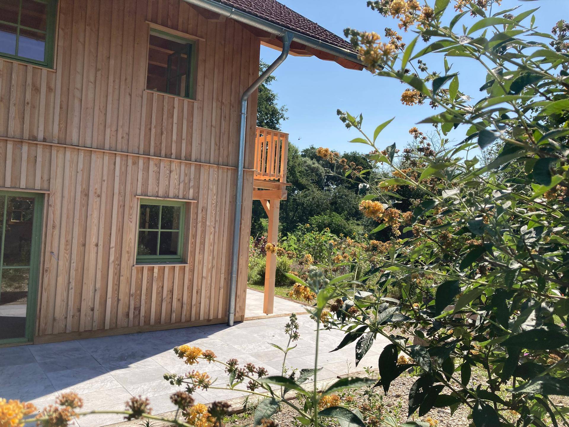 Seitenansicht des Holzhauses mit gepflastertem Bereich und blühenden Sträuchern im Garten.