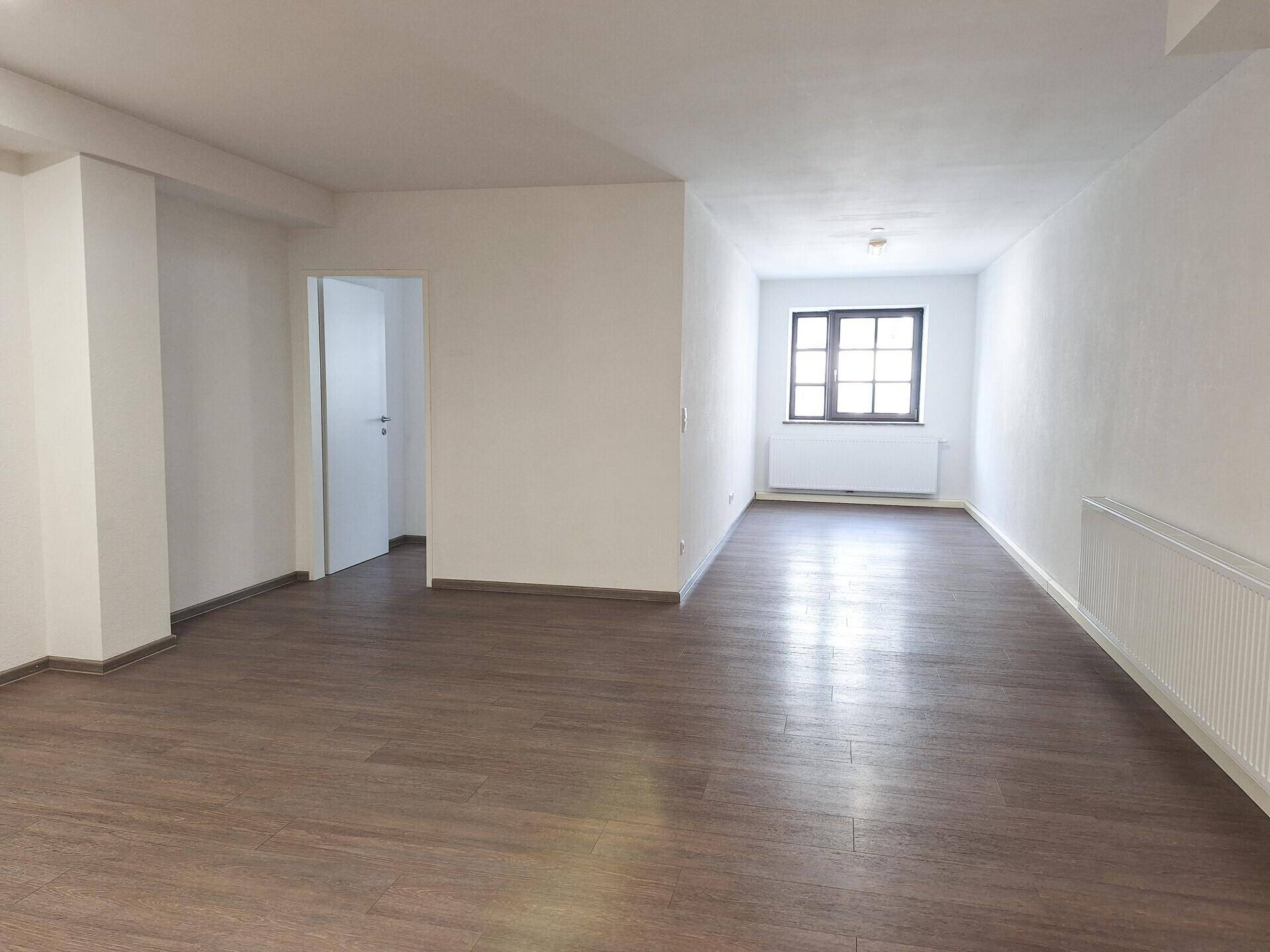 Geräumiger Wohnbereich mit Laminatboden, weißen Wänden und einem Fenster, das Tageslicht hereinlässt.