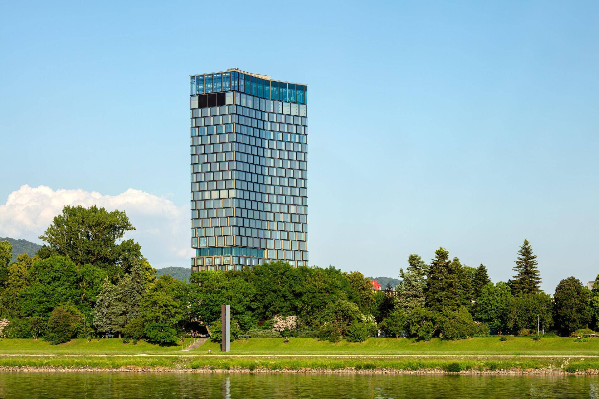 Hohes modernes Gebäude mit Glasfassade, umgeben von üppigem Grün und einem Fluss oder See im Vordergrund.