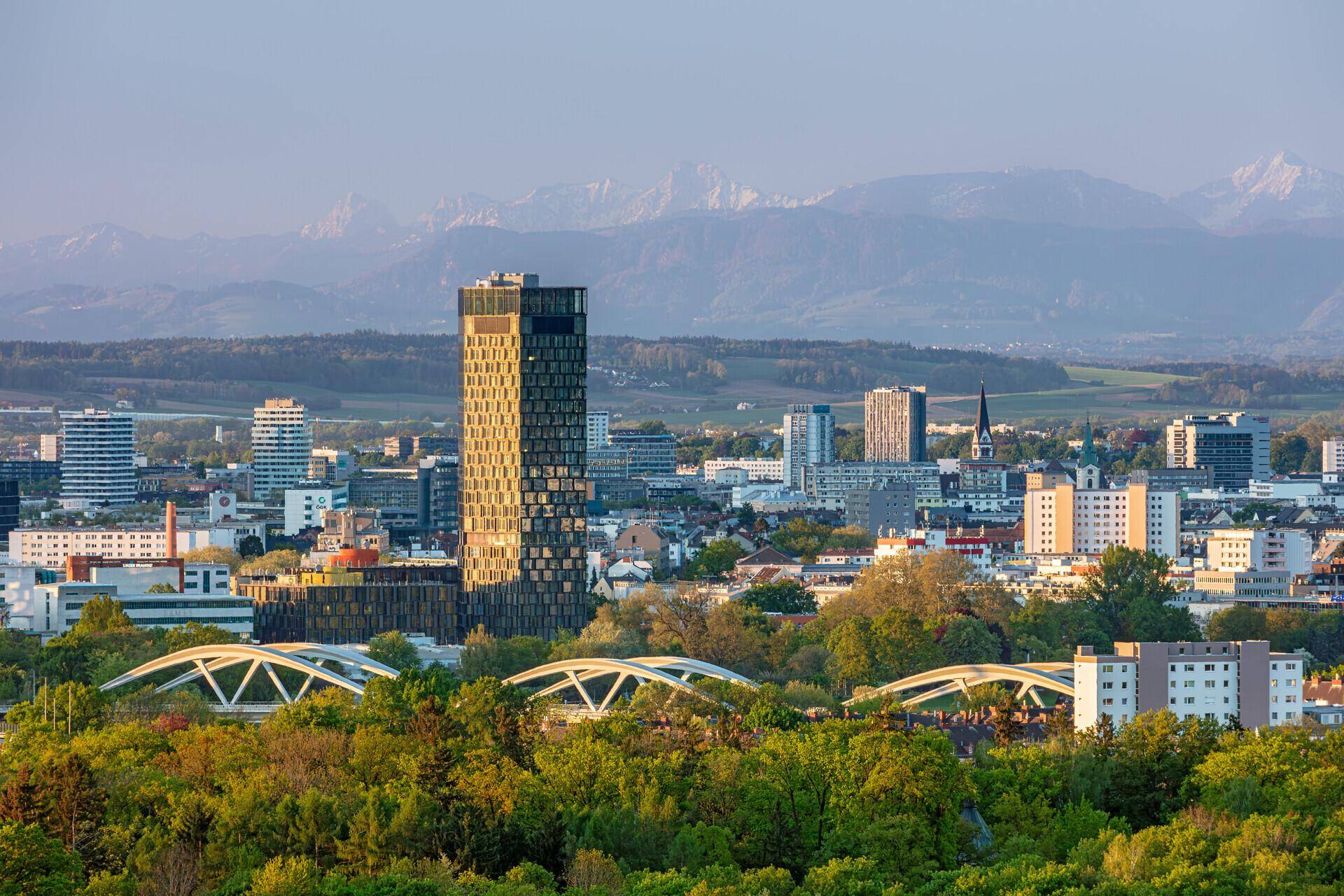 Panoramablick auf eine Stadtlandschaft mit einem markanten Hochhaus, grünen Brücken und schneebedeckten Bergen im Hintergrund.
