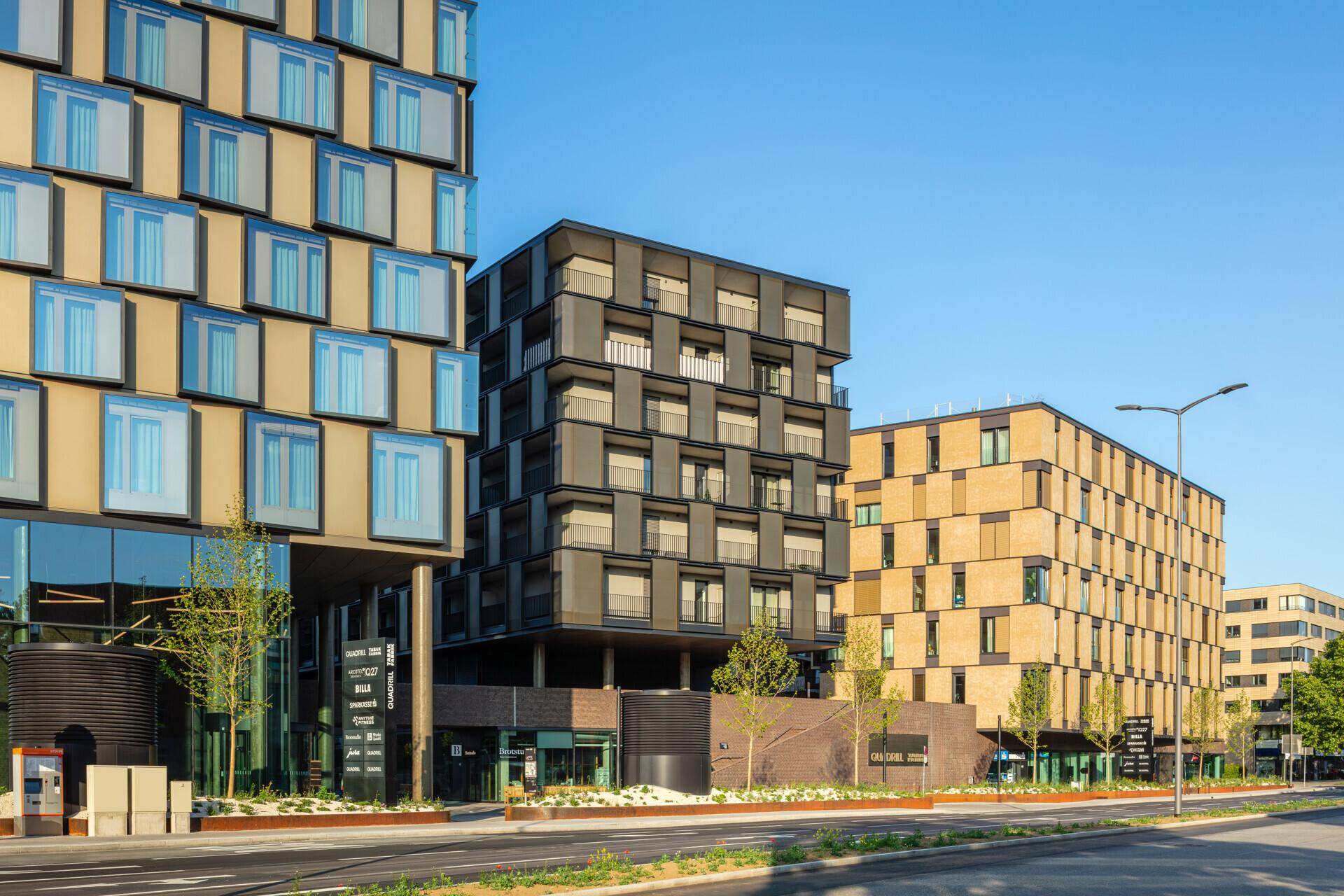 Moderne Gebäude mit markanten Fassaden und großen Fenstern entlang einer Straße, umgeben von Bäumen.