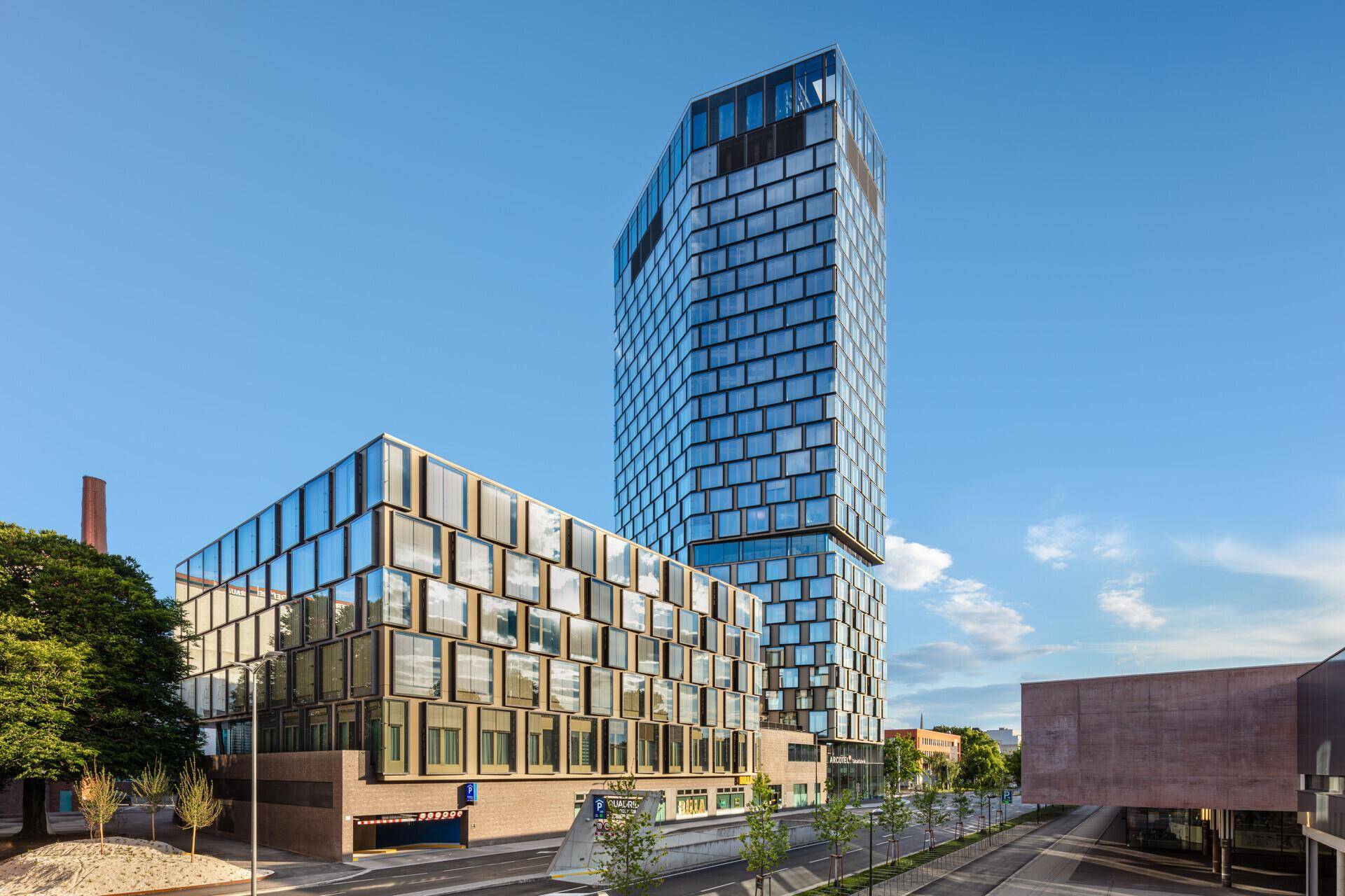 Zwei moderne Gebäude mit Glasfassaden und markanter Architektur, eines hoch aufragend, das andere niedriger, an einer Straße.