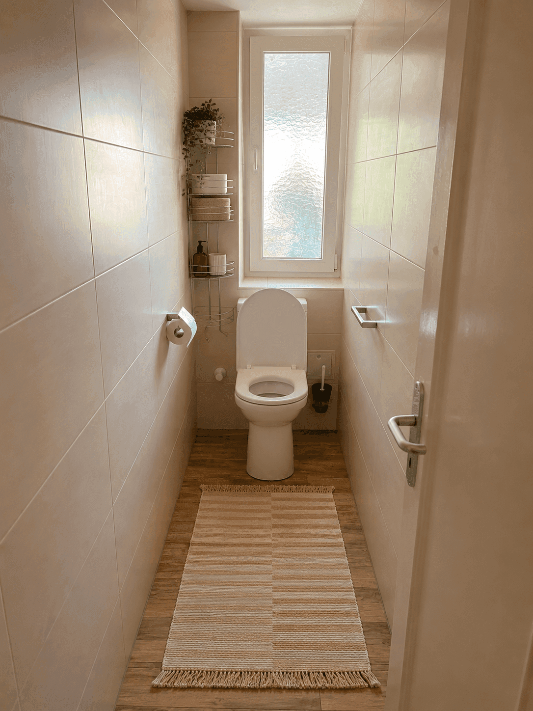 Kleines WC mit Toilette, Waschbecken und Fenster, gefliesten Wänden und Holzoptikboden.
