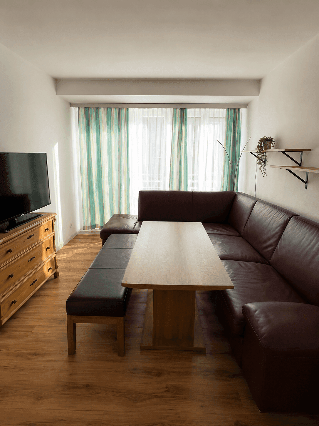 Helles Wohnzimmer mit großem Ledersofa, Holztisch und Fernseher vor gestreiften Vorhängen.