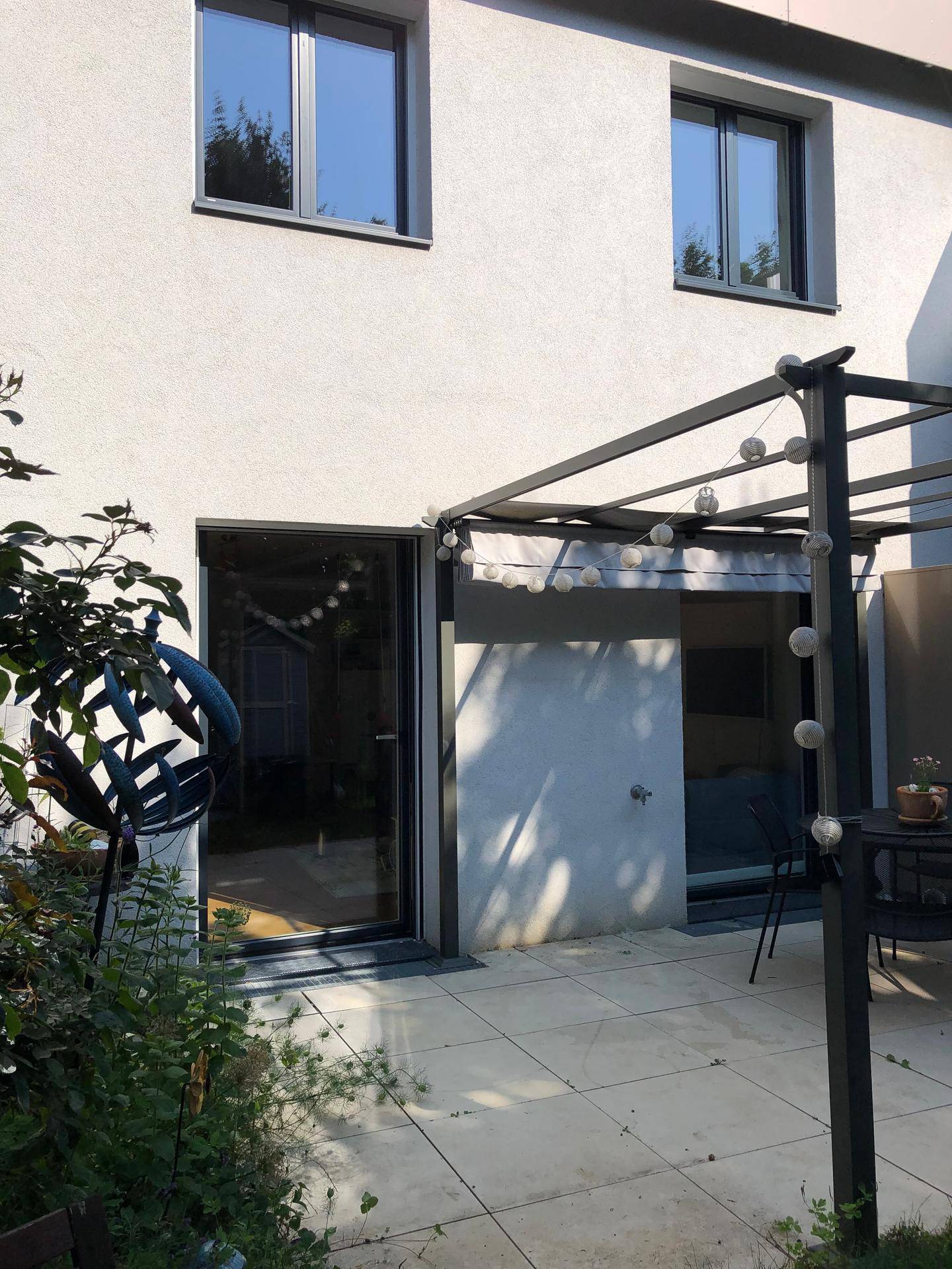 Helle Terrasse mit Pergola und Lichterkette vor der modernen Hausfassade.