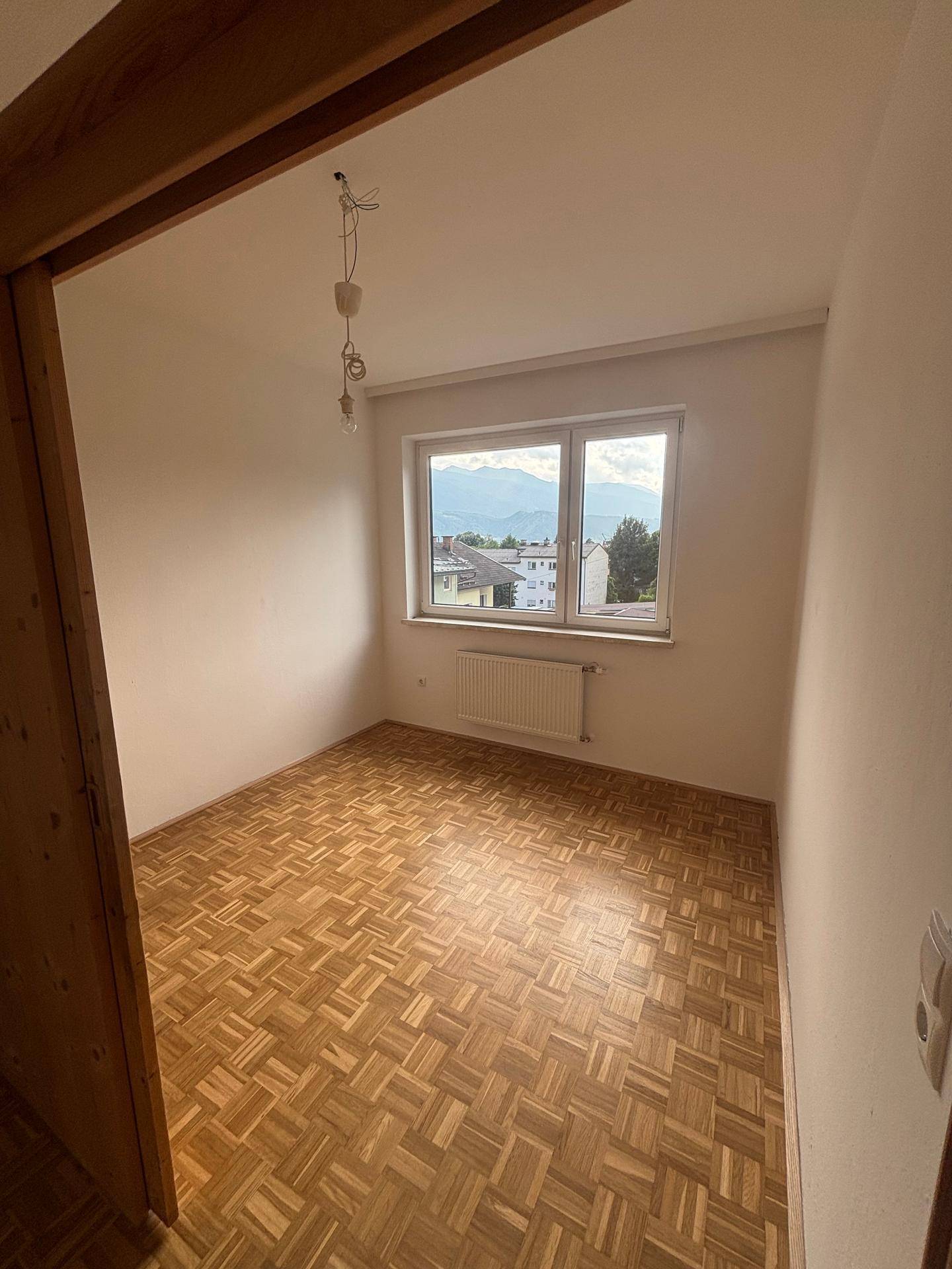 Leeres Zimmer mit klassischem Parkettboden, weißen Wänden und einem Fenster mit Bergblick.