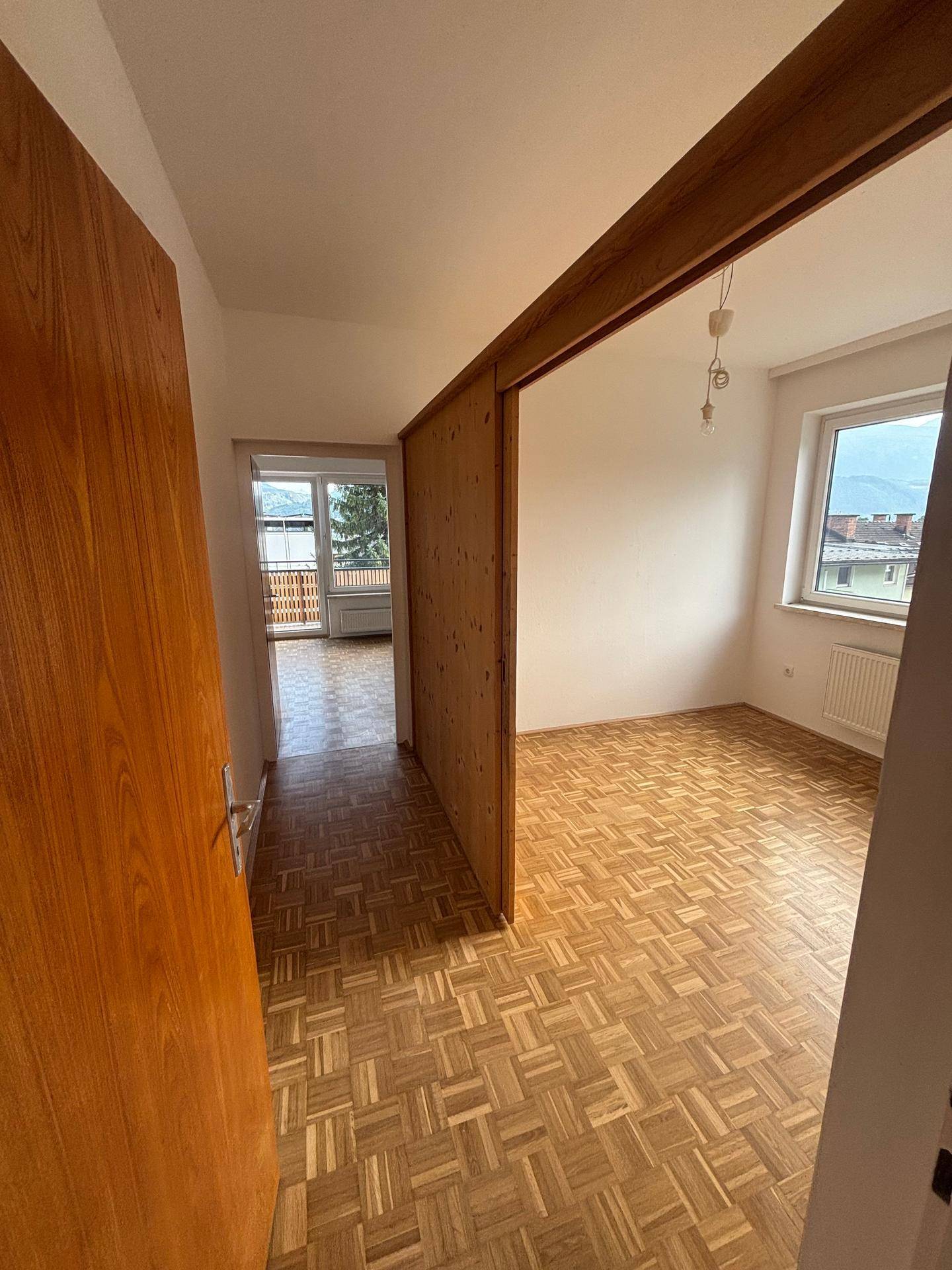 Blick aus einem Flur in ein Zimmer mit Parkettboden und weiter auf einen Balkon mit Bergblick.