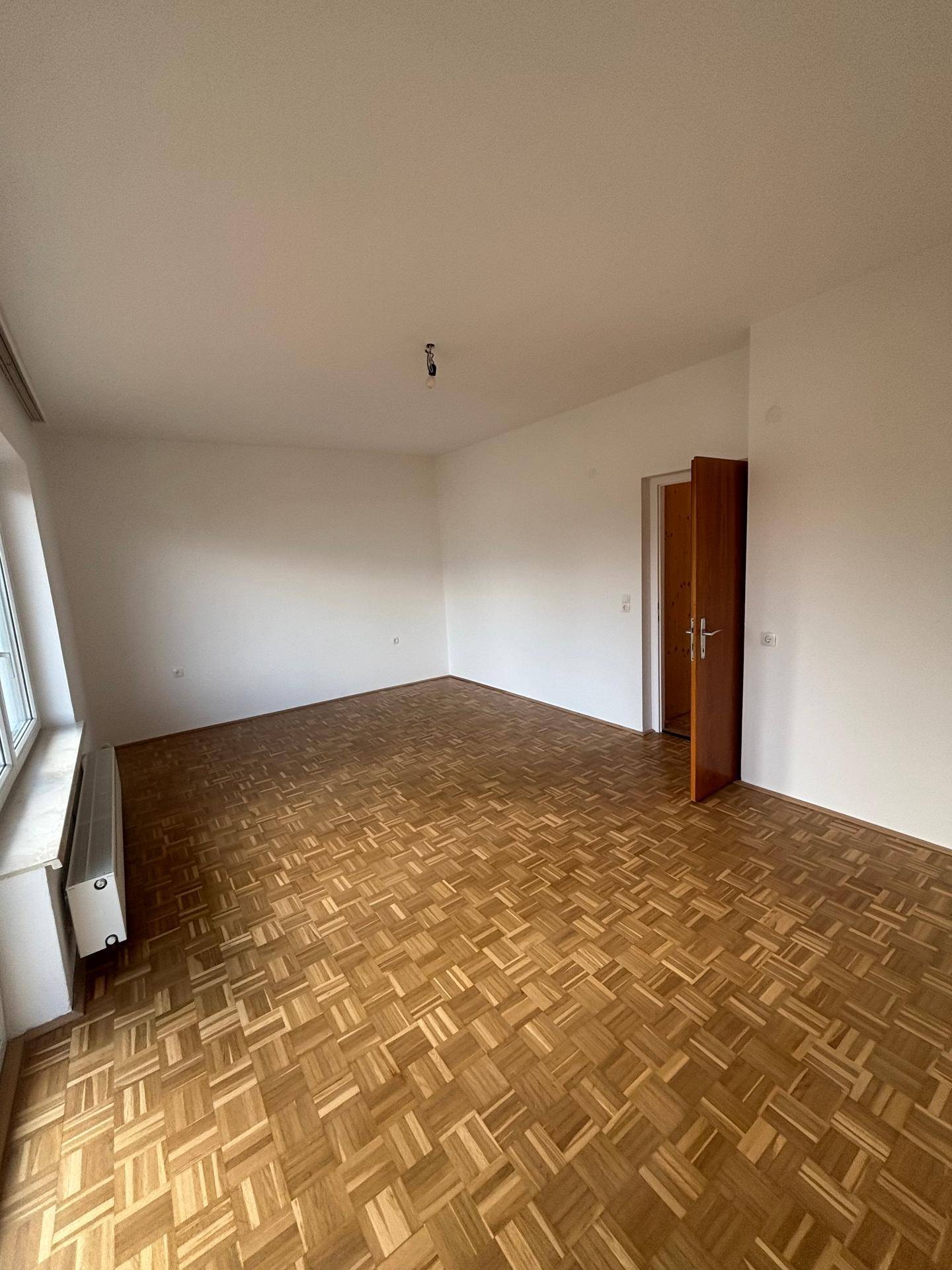 Geräumiges, leeres Zimmer mit Parkettboden, weißen Wänden und viel natürlichem Licht durch das Fenster.