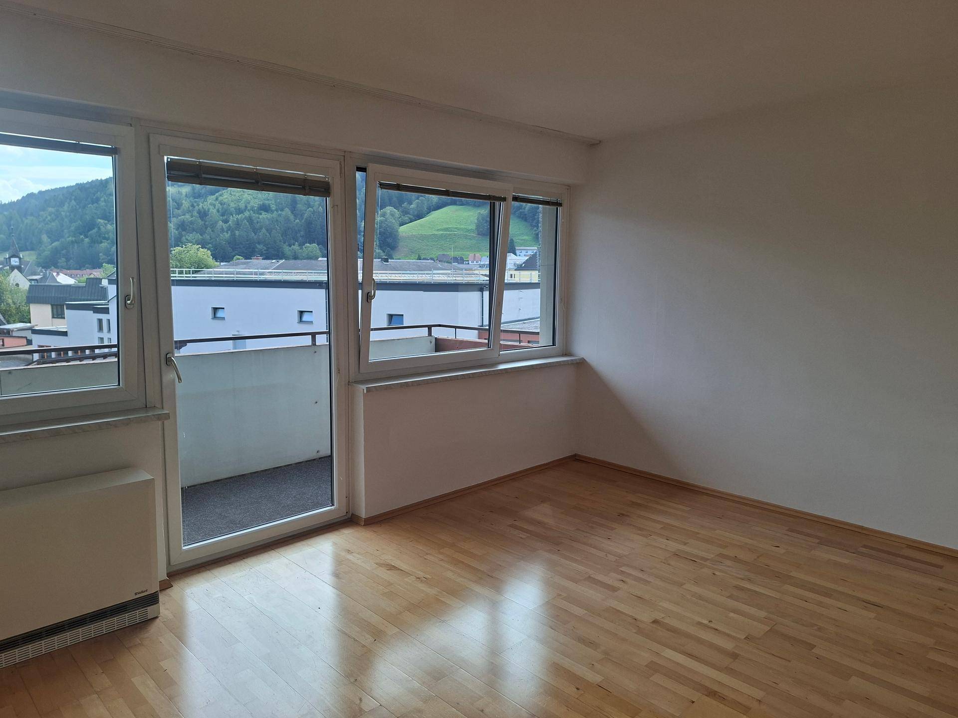 Helles Zimmer mit Holzboden, großem Fenster und Zugang zu einem Balkon mit Bergblick.