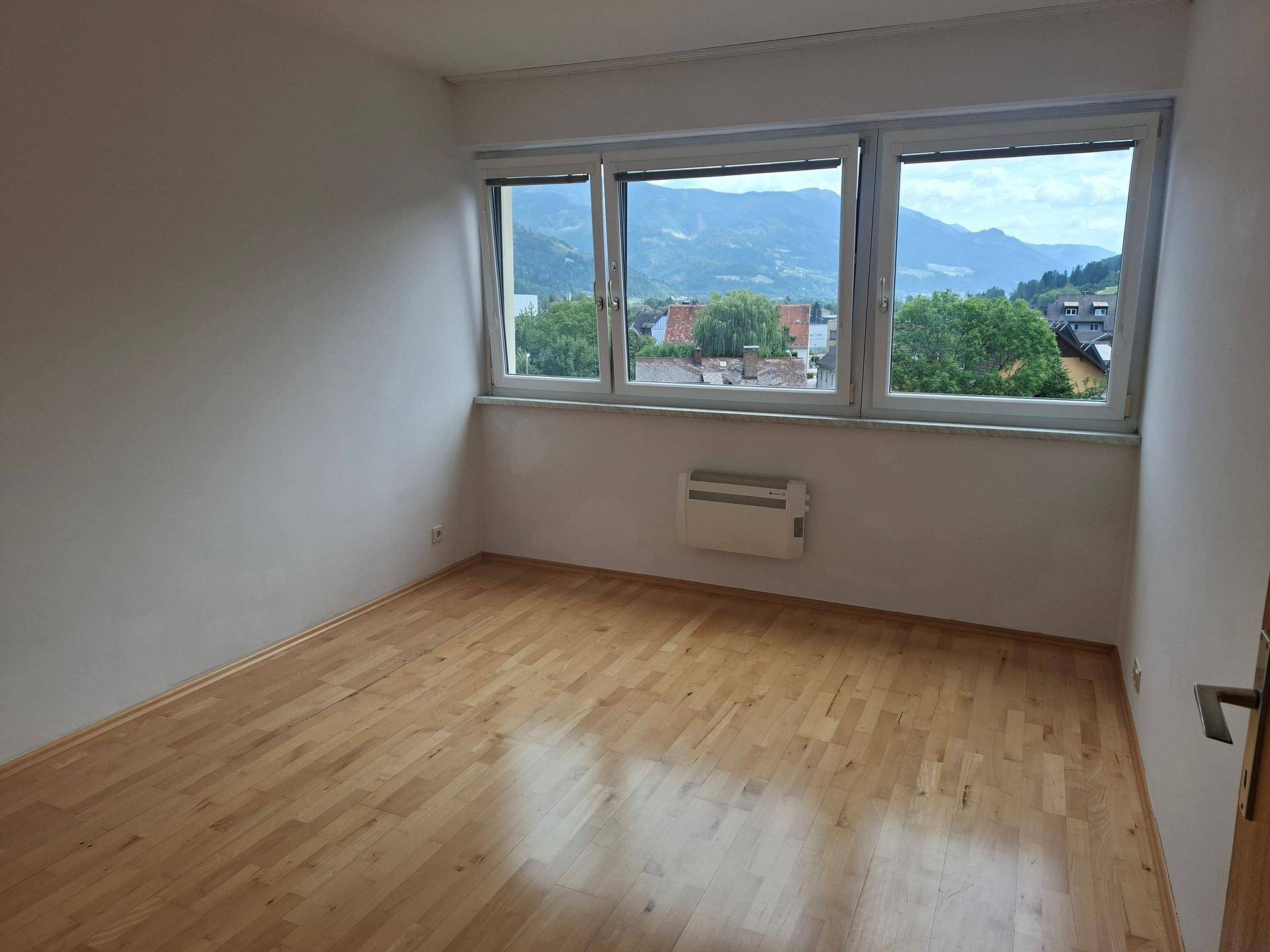 Geräumiges Zimmer mit Holzboden und breitem Fenster mit weitem Blick auf die Berge.