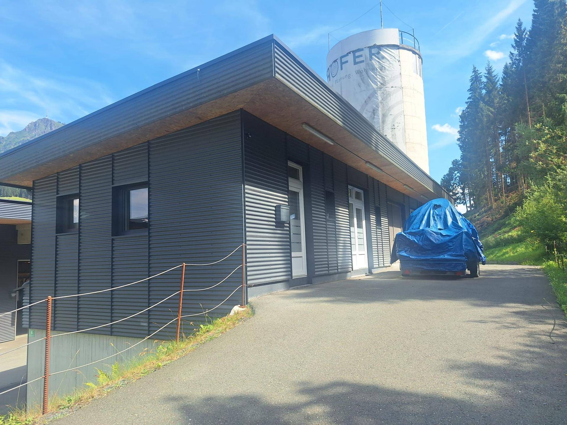 Modernes Gebäude mit geriffelter Fassade und einem Wasserturm im Hintergrund, umgeben von Bäumen.