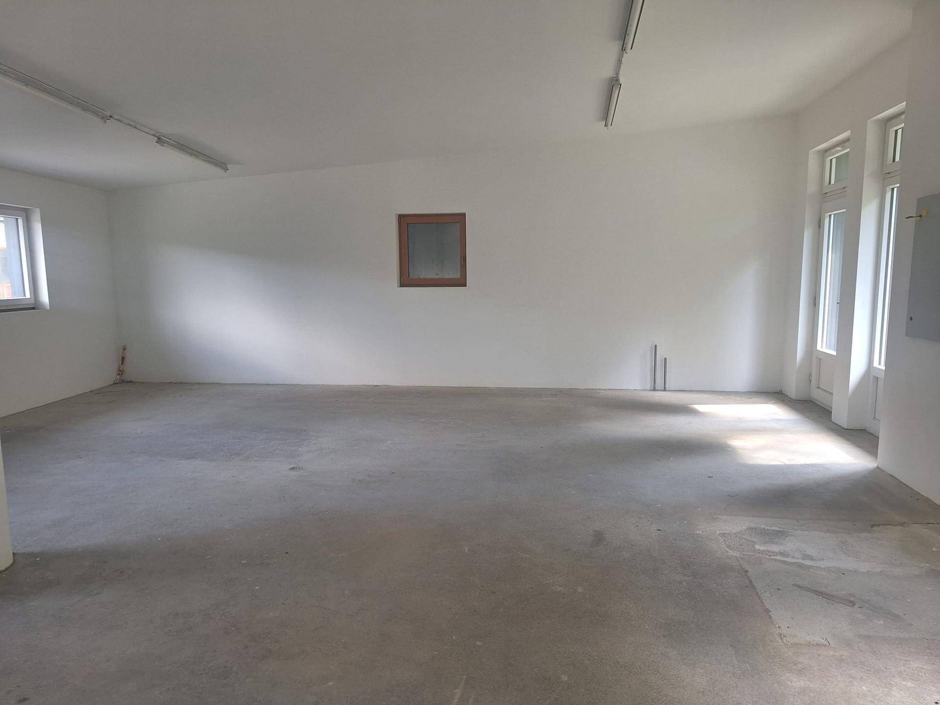 Großer, leerer Raum mit Betonboden, weißen Wänden und mehreren Fenstern, ideal für gewerbliche Nutzung.
