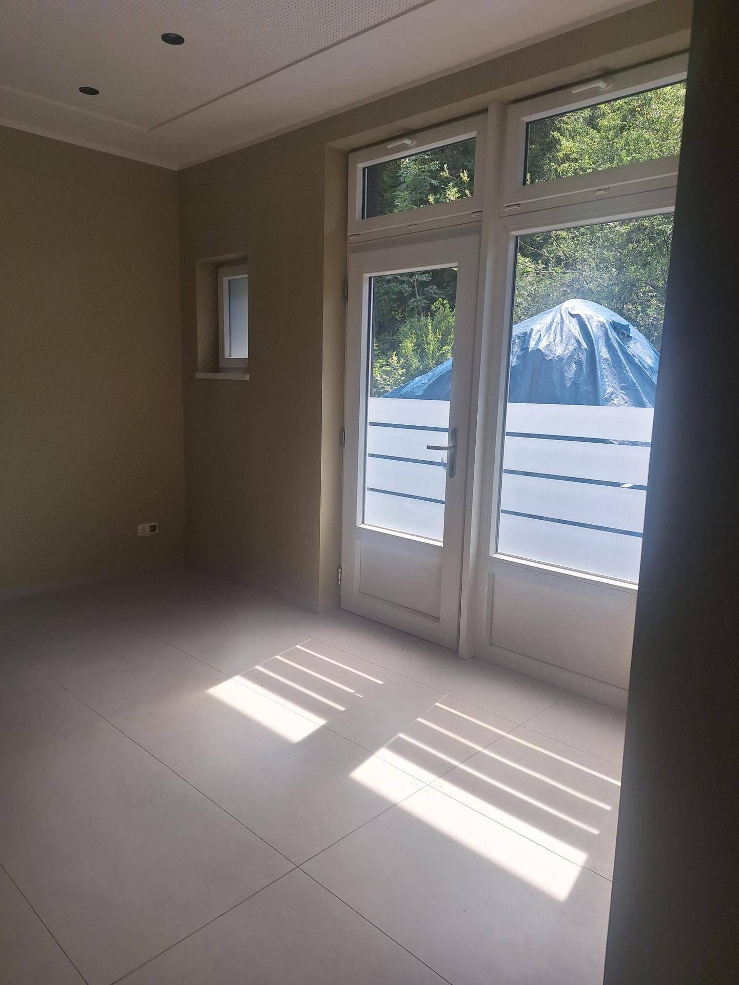 Heller, leerer Raum mit Fliesenboden und großen Fenstertüren, die viel Tageslicht hereinlassen.