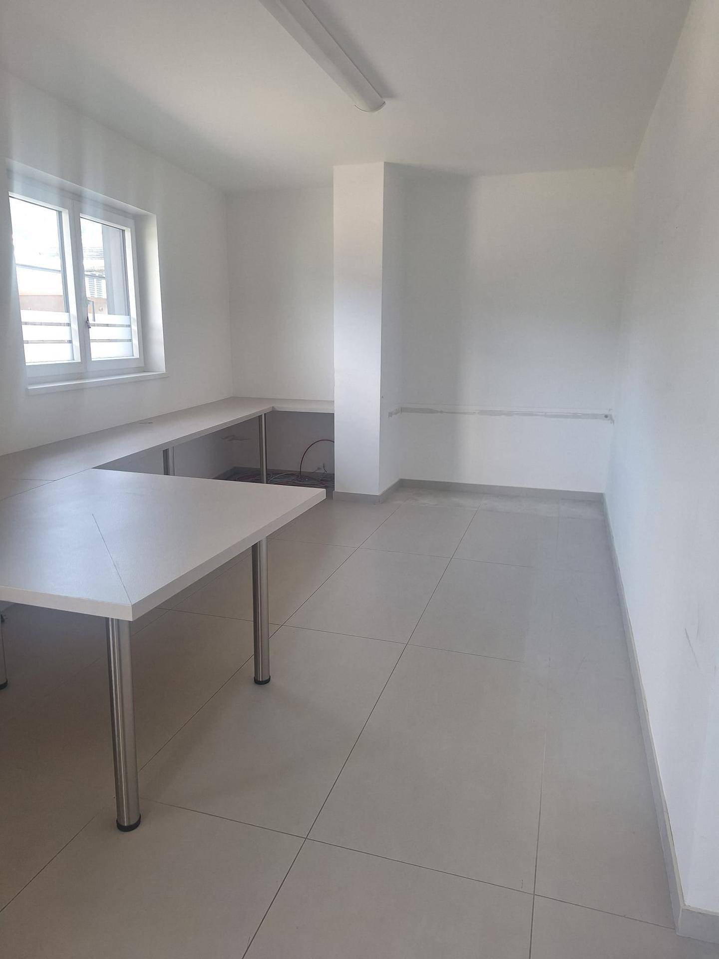 Funktionaler Büroraum mit weißen Wänden, Fliesenboden und langen Arbeitstischen entlang der Wand.