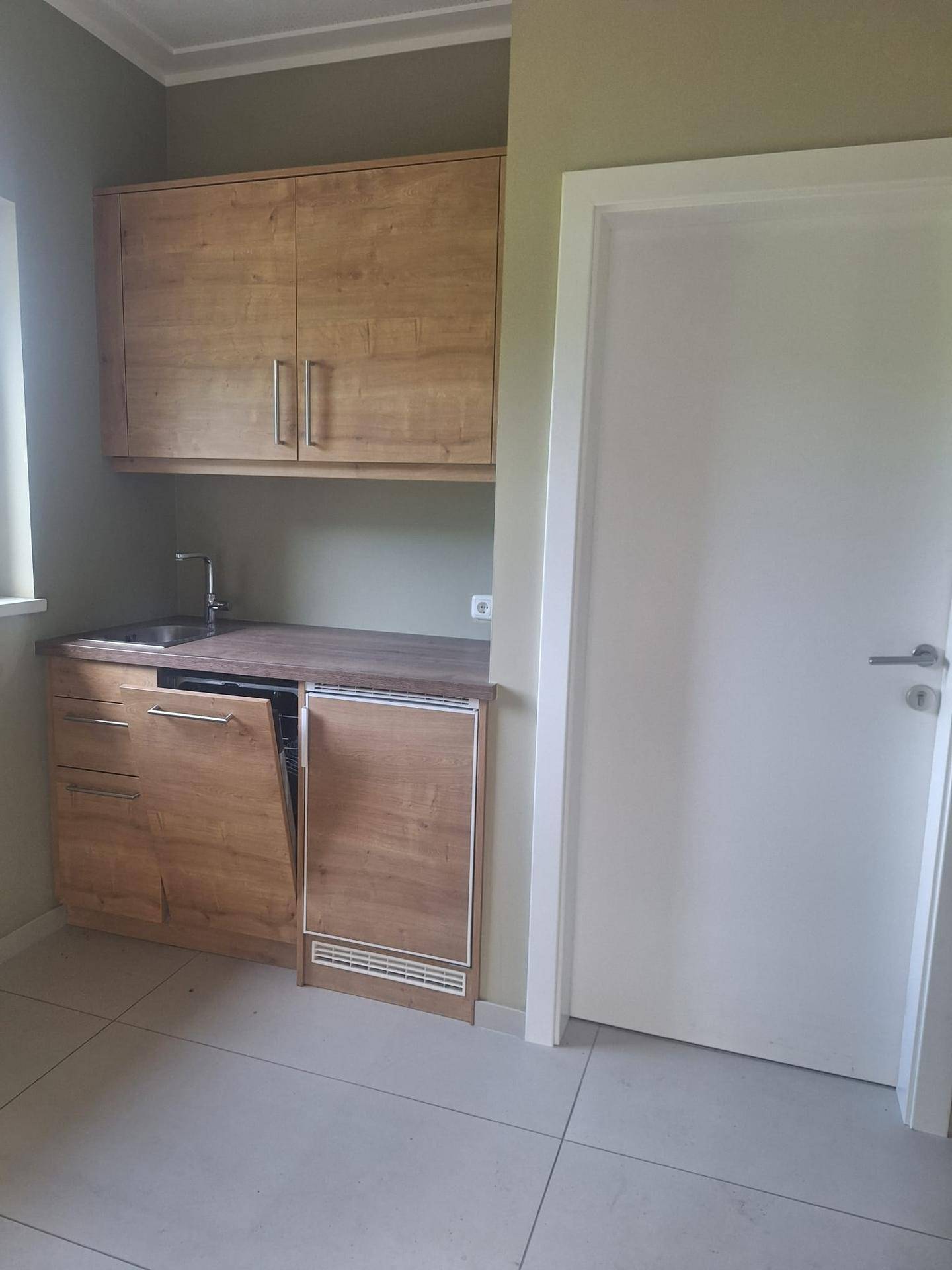 Kleine Küchenzeile mit Holzschränken, Spüle und integriertem Kühlschrank in einem hellen Raum.