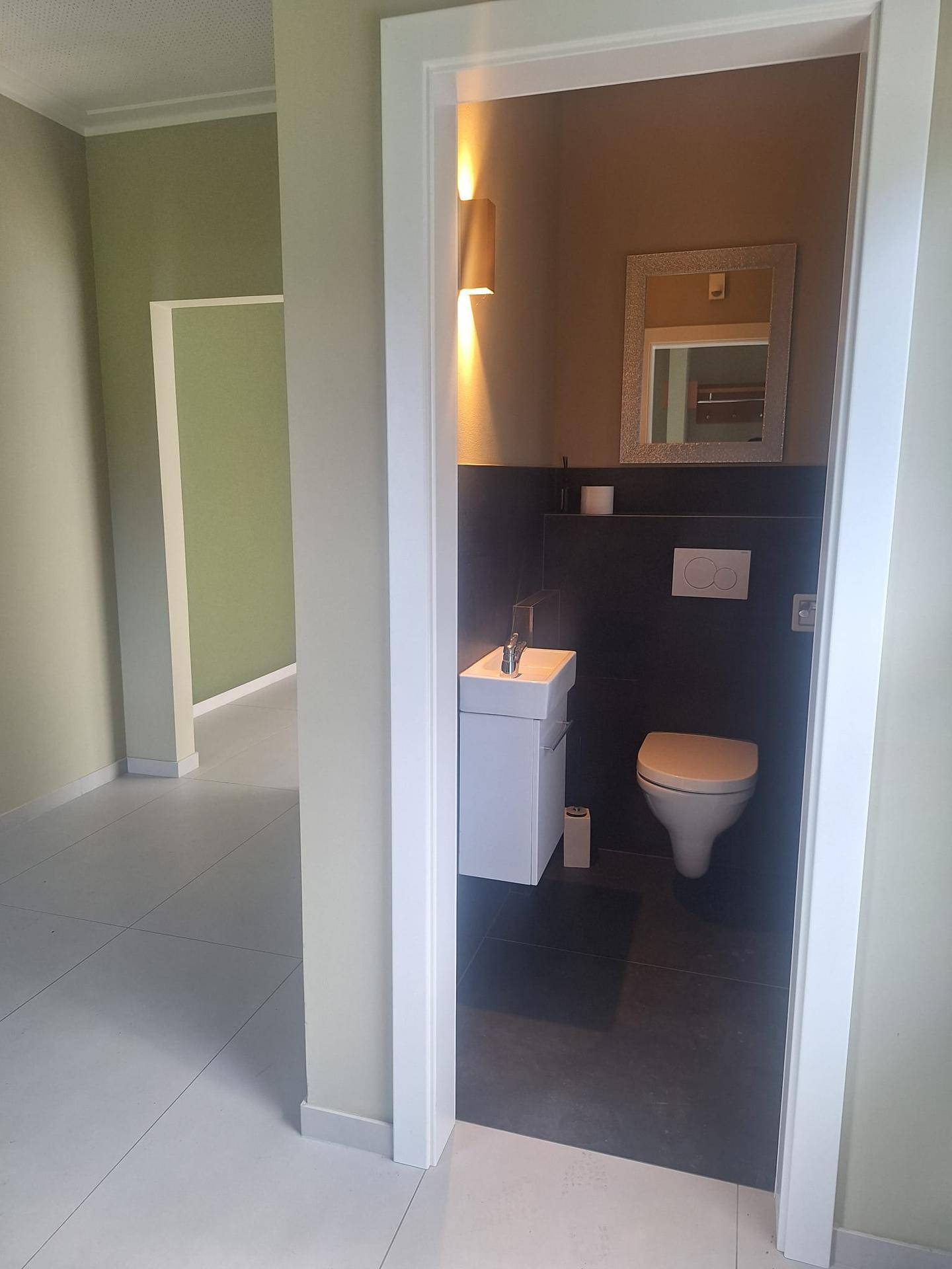 Modernes Gäste-WC mit dunklen Fliesen, wandhängendem WC und kleinem Waschbecken mit Spiegel.
