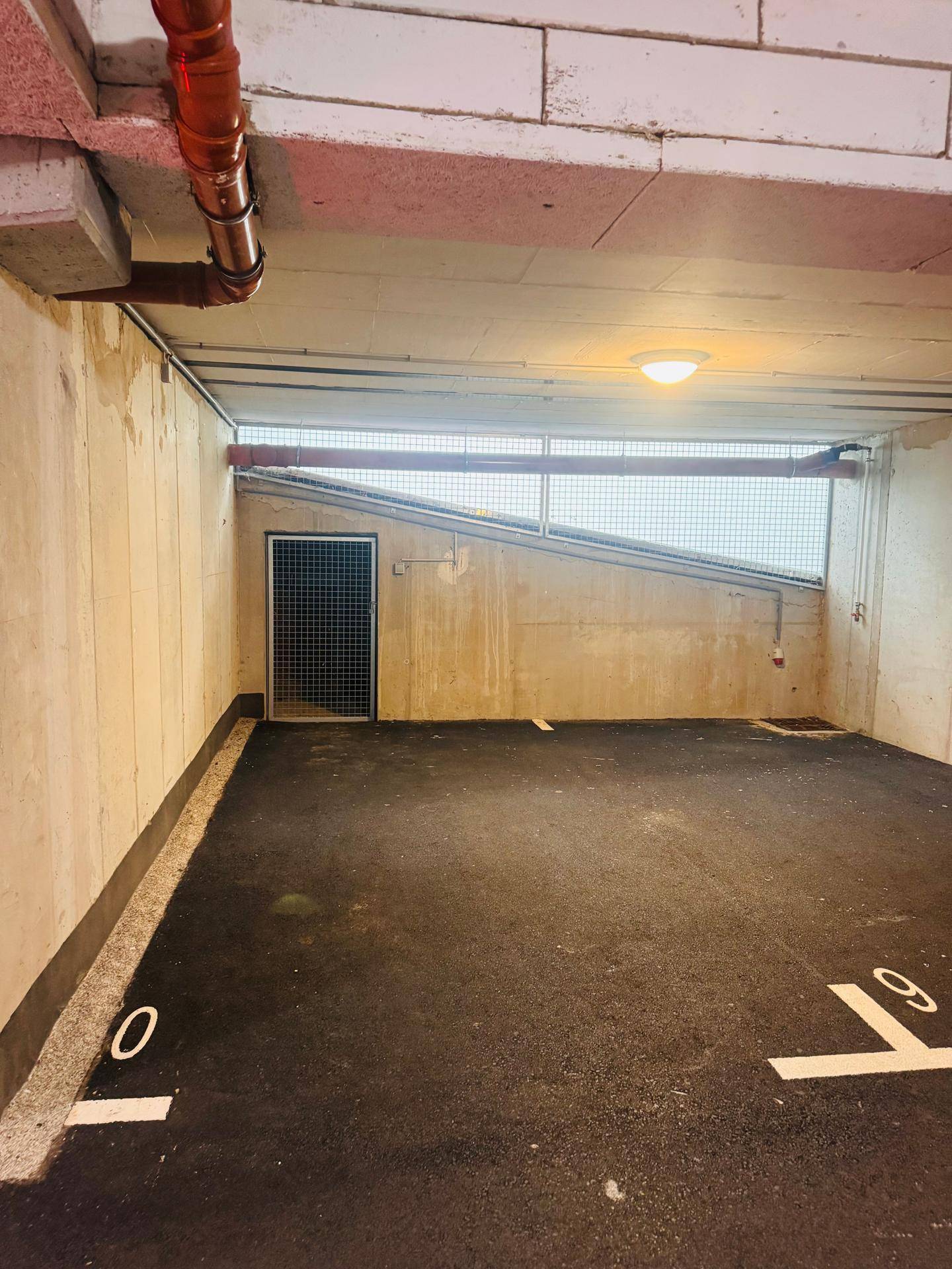 Tiefgaragenstellplatz mit sichtbaren Rohren an der Decke und Gittertür zu einem Abstellraum.