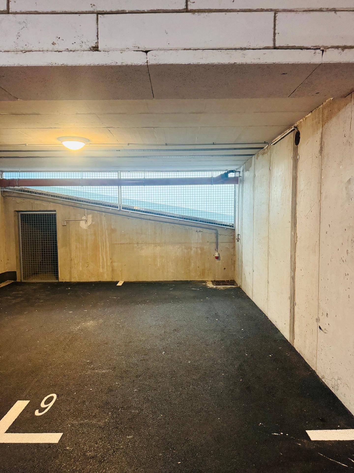 Leerer Parkplatz in einer Tiefgarage mit Betonwänden und Gittertür zu einem Abstellraum.