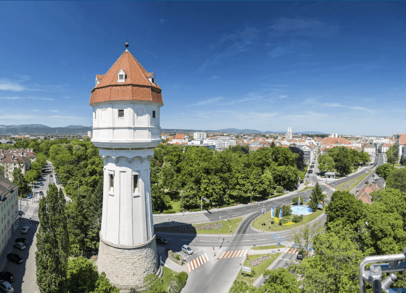 Panoramablick auf die Stadt mit einem markanten Wasserturm, Straßen und Grünflächen bei Tag.